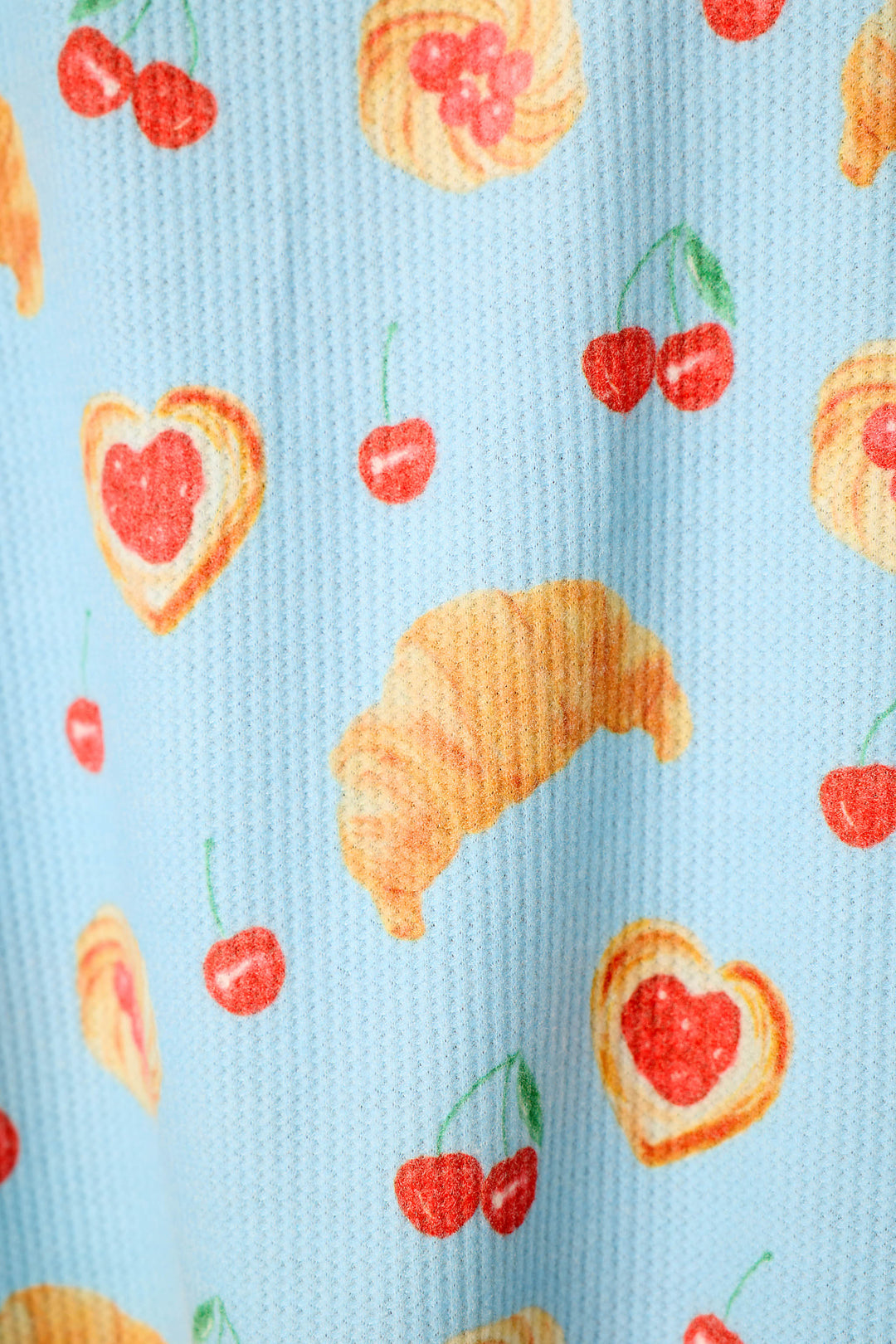A close-up of a light blue textured fabric printed with a repeating pattern of red cherries and croissants, including heart-shaped croissants with red jam centers.