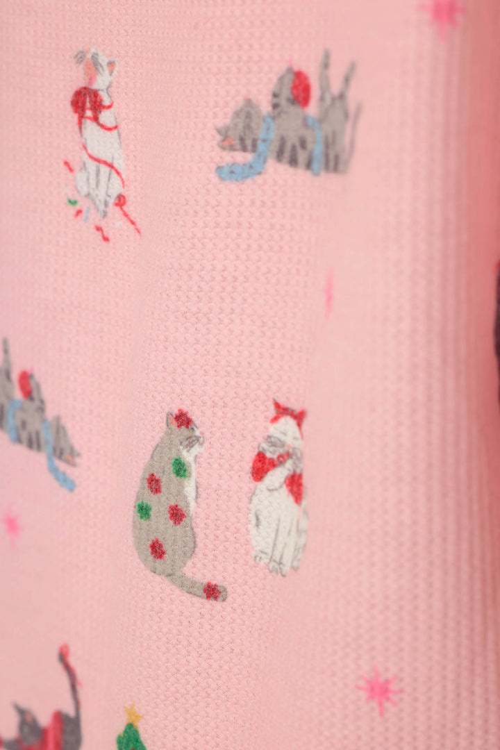 A close-up of a pink textured fabric featuring a whimsical print of cats dressed in winter clothing, including scarves, and green and red bows.