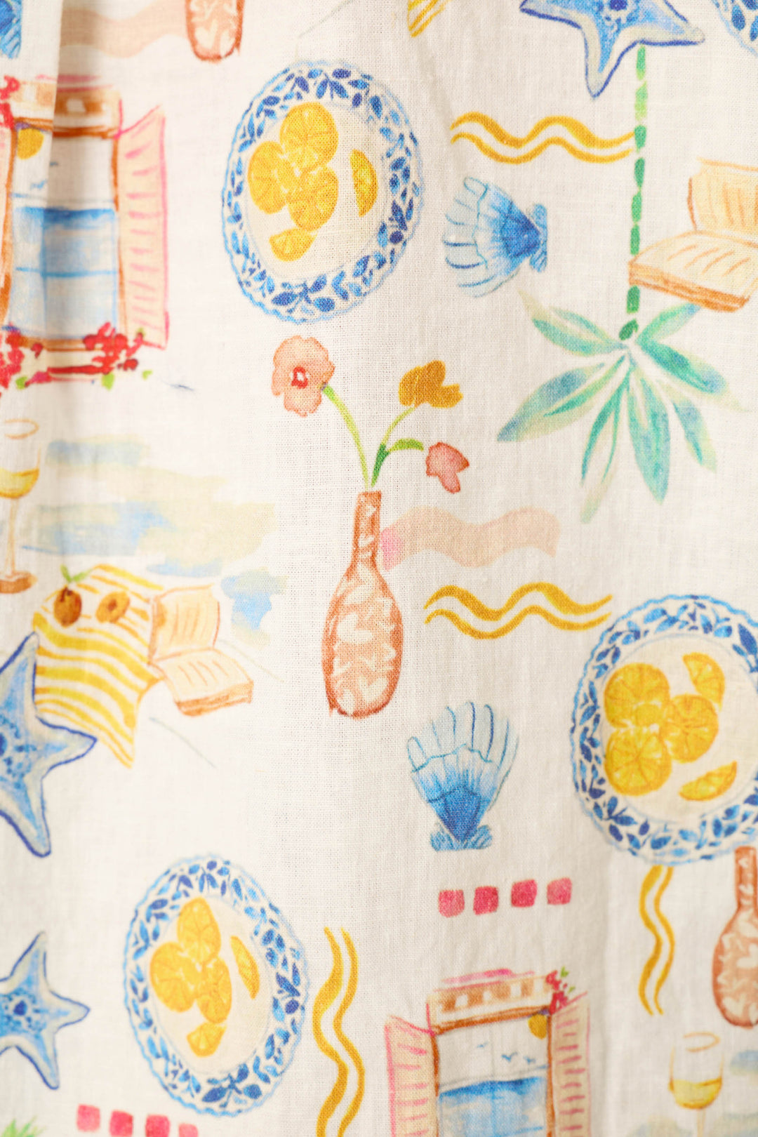 White fabric with a vibrant print of plates, lemons, seashells, flowers in vases, starfish, palm leaves, books, and wine glasses.