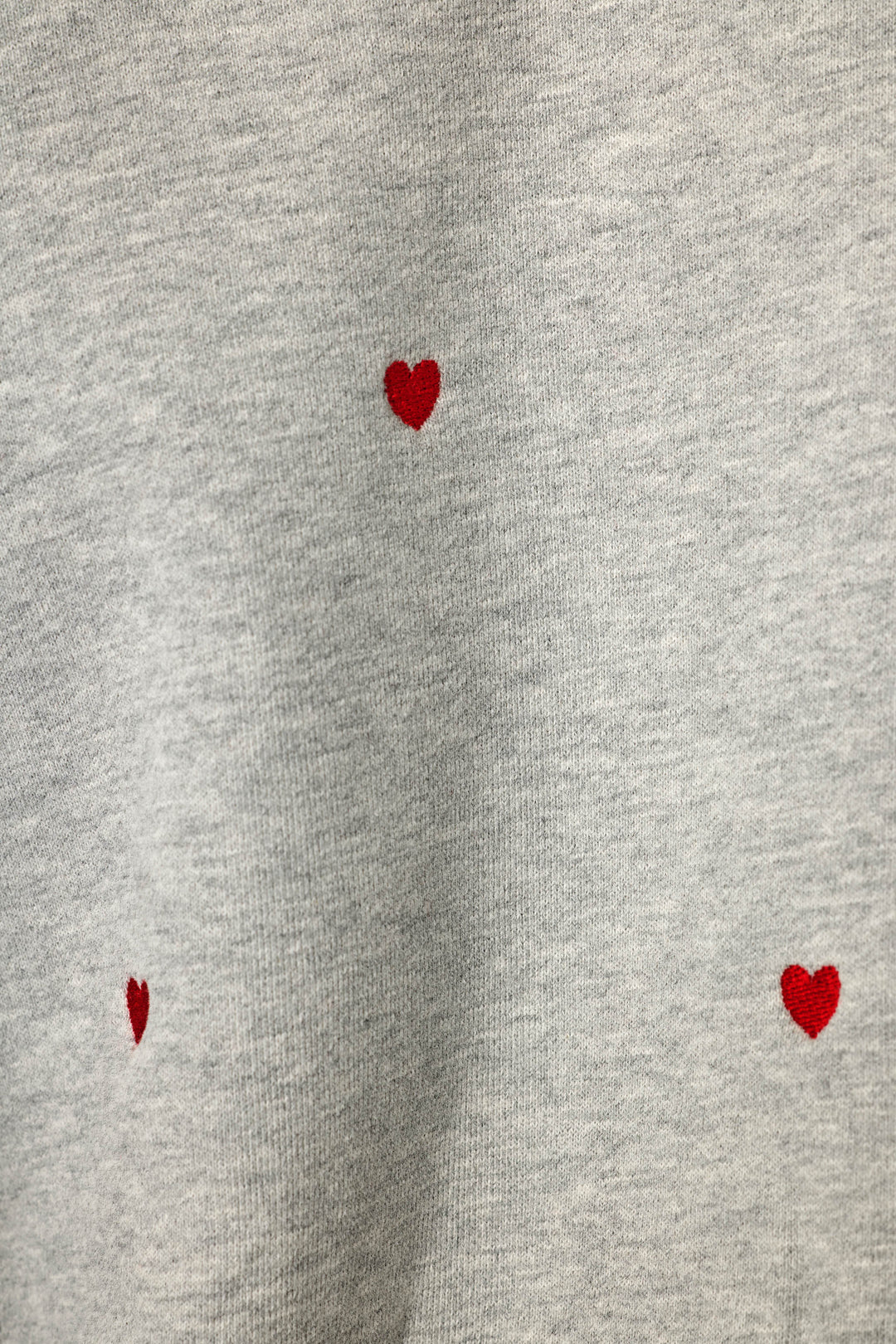 Close-up of a light heather gray sweatshirt style fabric with small red hearts embroidered into the fabric and scattered across it, widely spaced apart.