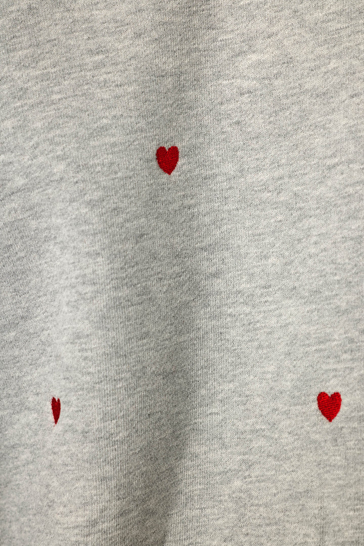 Close-up of a light heather gray sweatshirt style fabric with small red hearts embroidered into the fabric and scattered across it, widely spaced apart.