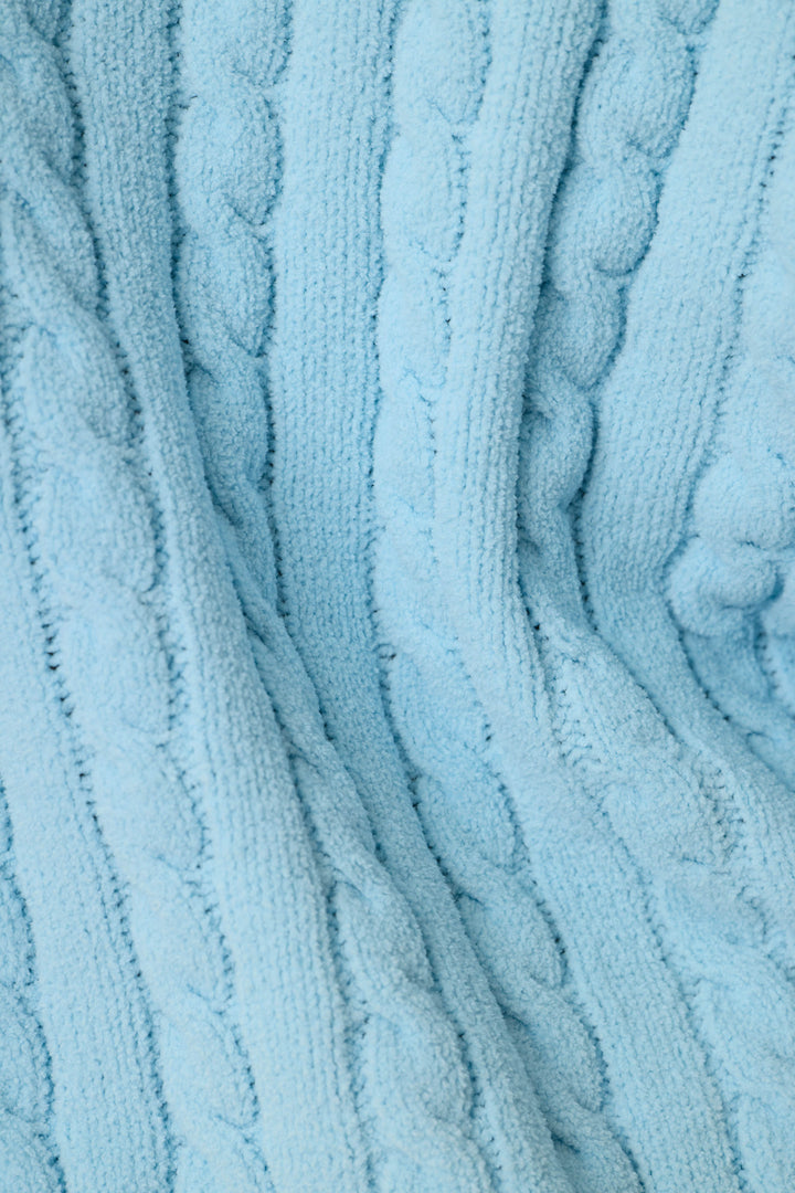 A close-up of a light blue cable-knit fabric texture, showing the detailed knit pattern and soft folds.