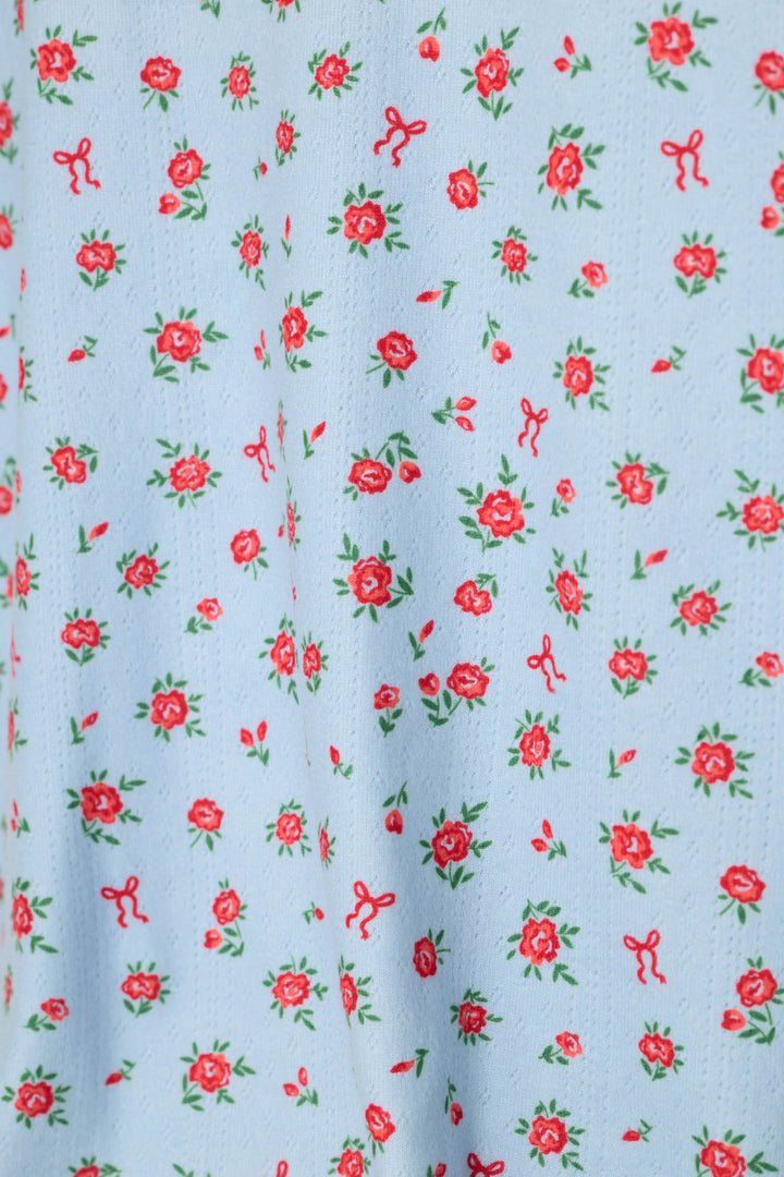 A close-up of a light blue fabric with a repeating pattern of small red roses, bows, and green leaves.