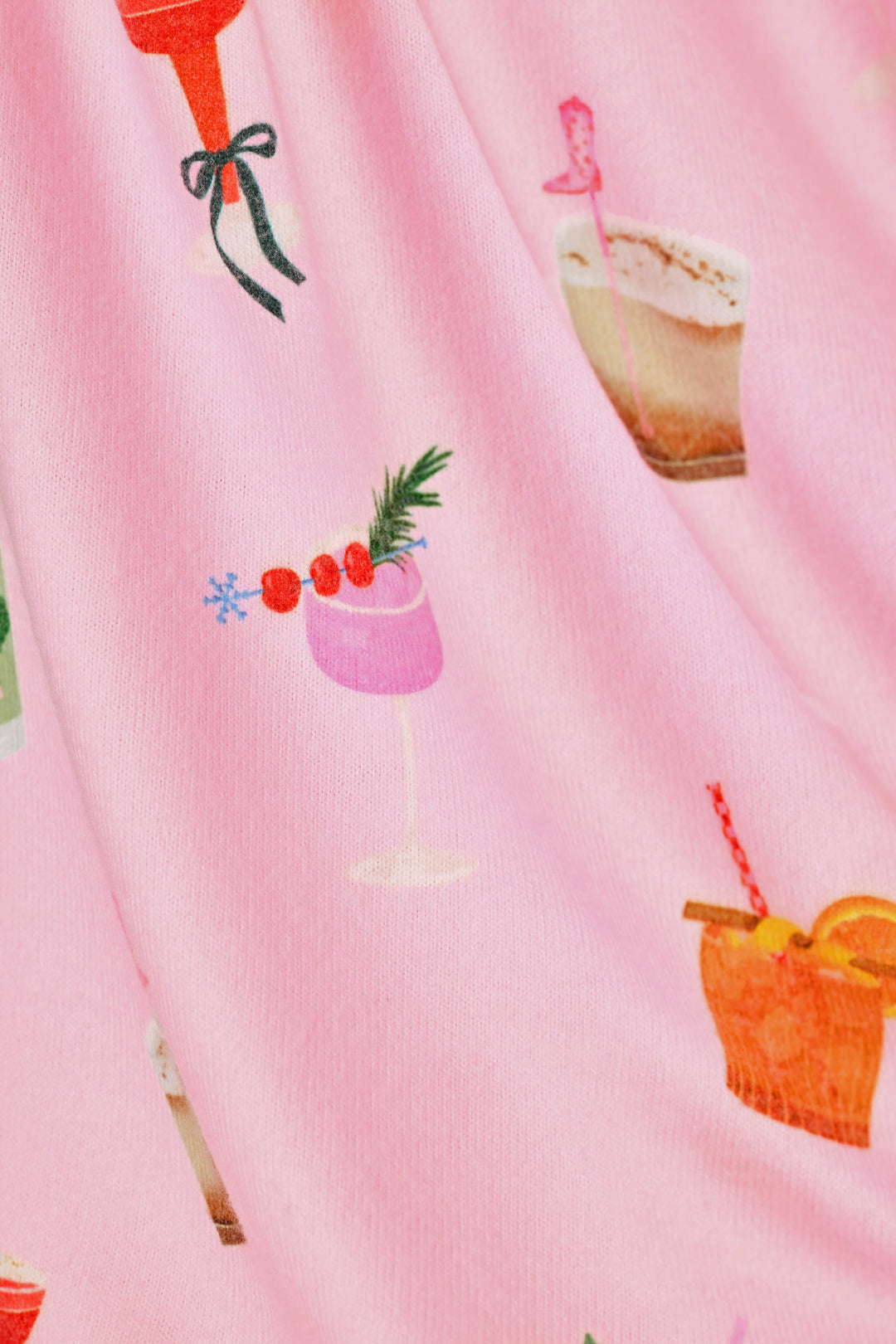 A close-up of a pink fabric with a festive cocktail print, showing illustrations of various drinks garnished with cherries, pine needles, and straws.