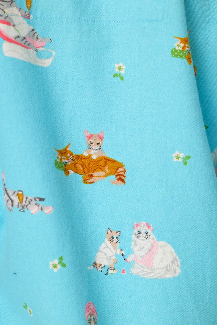 A close-up of a sky blue fabric printed with cute illustrated cats in orange, white, and gray enjoying a day at the spa, and also has small white flowers sprinkled  throughout.