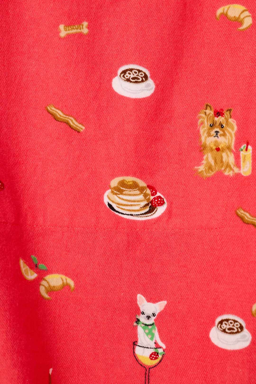 A close-up of a dark coral fabric with a whimsical, printed pattern featuring pancakes, croissants, coffee cups, bacon strips, and small dogs ready for brunch.