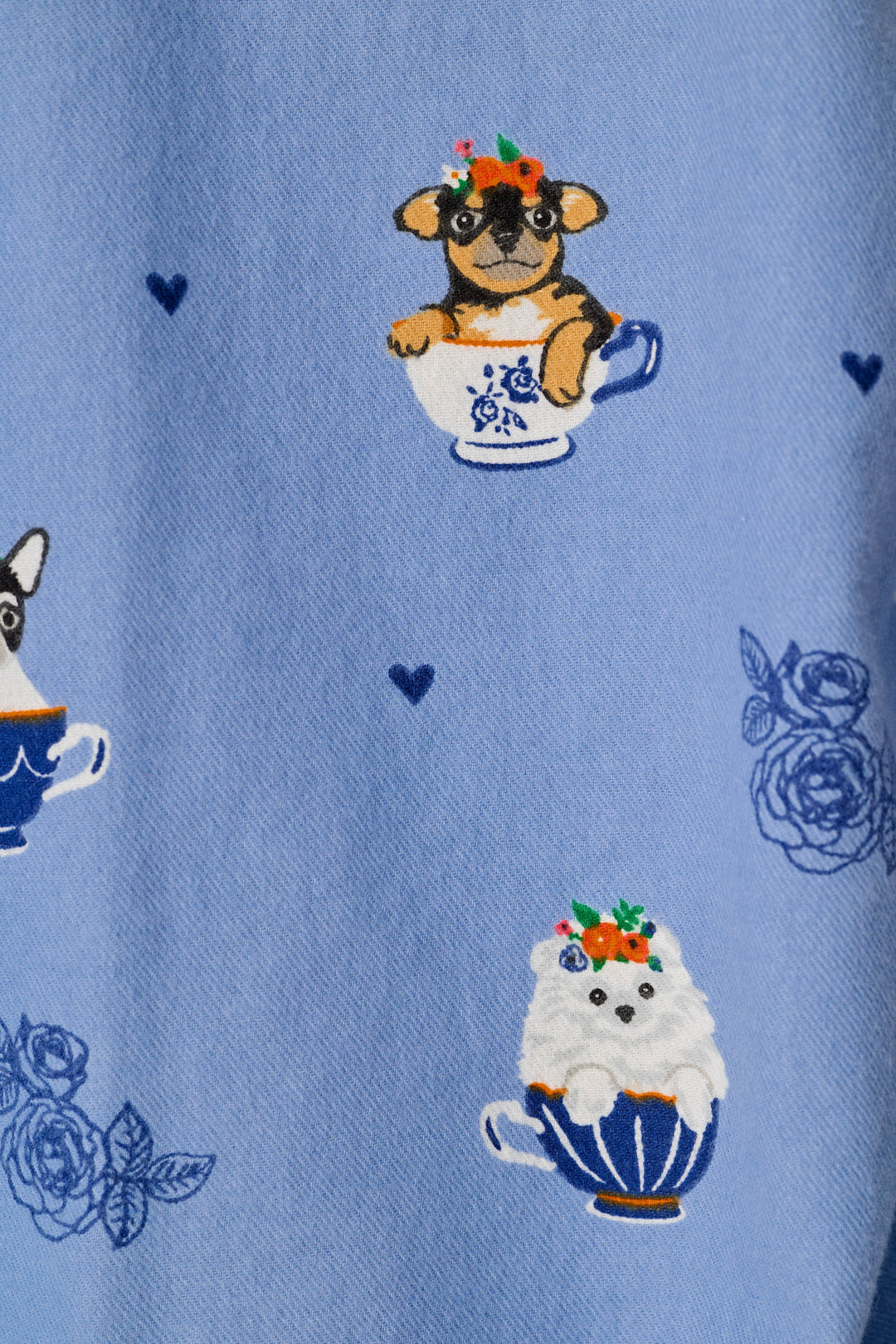 A close-up of a light blue fabric featuring a whimsical pattern of small dogs sitting in teacups with floral crowns, alongside blue rose motifs and tiny heart details.