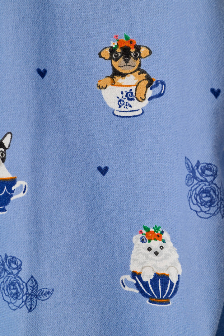 A close-up of a light blue fabric featuring a whimsical pattern of small dogs sitting in teacups with floral crowns, alongside blue rose motifs and tiny heart details.