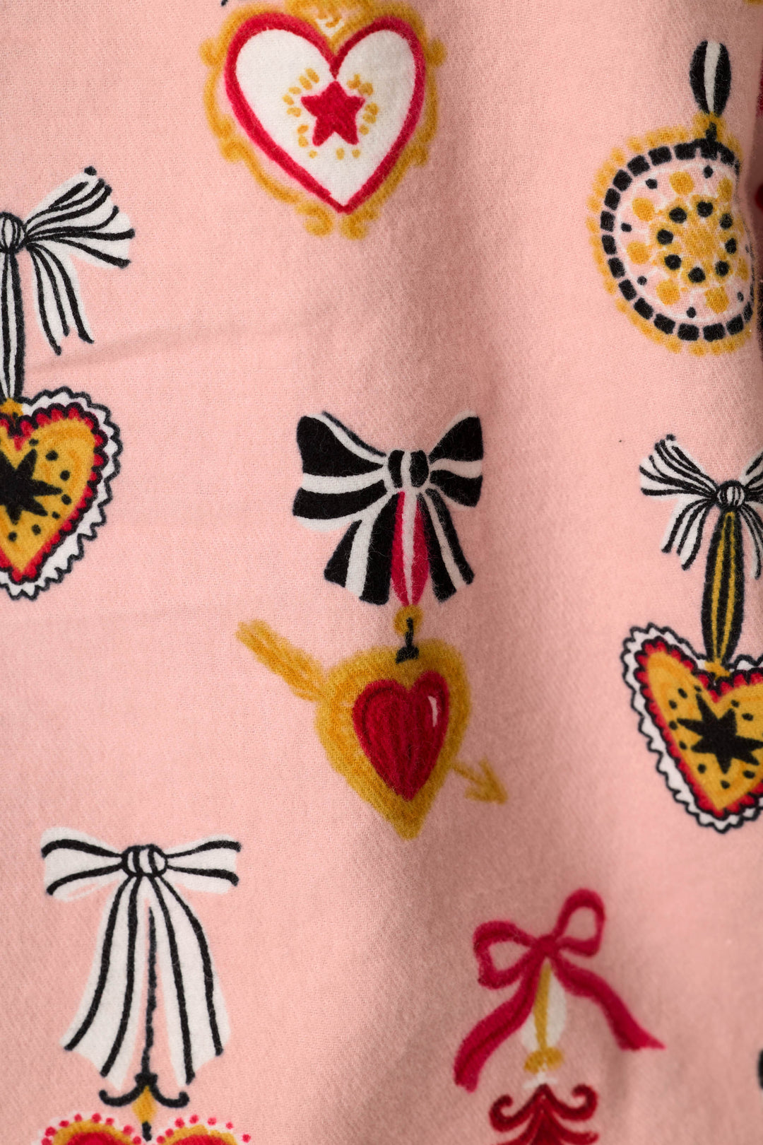 A close-up of a light pink fabric featuring a repeating pattern of stylized hearts, bows, and decorative motifs in red, yellow, black, and white.