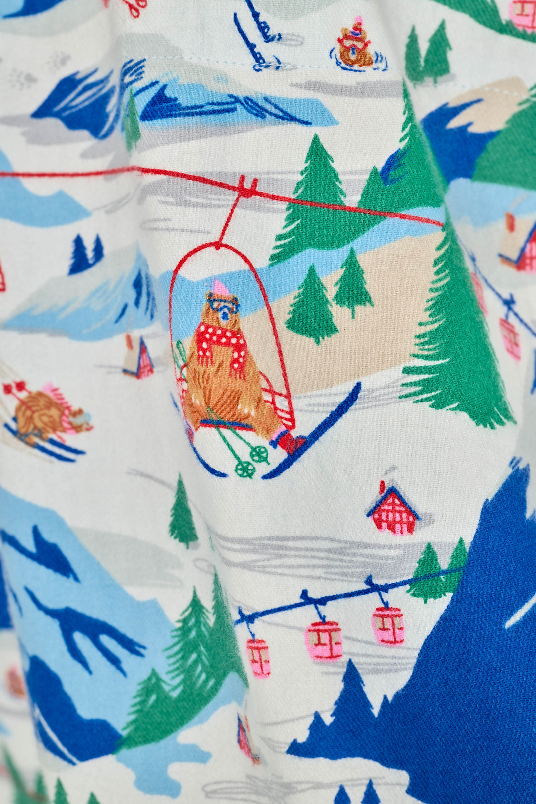 A close-up of a fabric with a whimsical winter mountain print, featuring a brown bear in a red polka-dot ski suit riding a red cable car, with blue mountains, green trees, and red cabins on a white background. Also featuring bears skiing down a snowy mountain.
