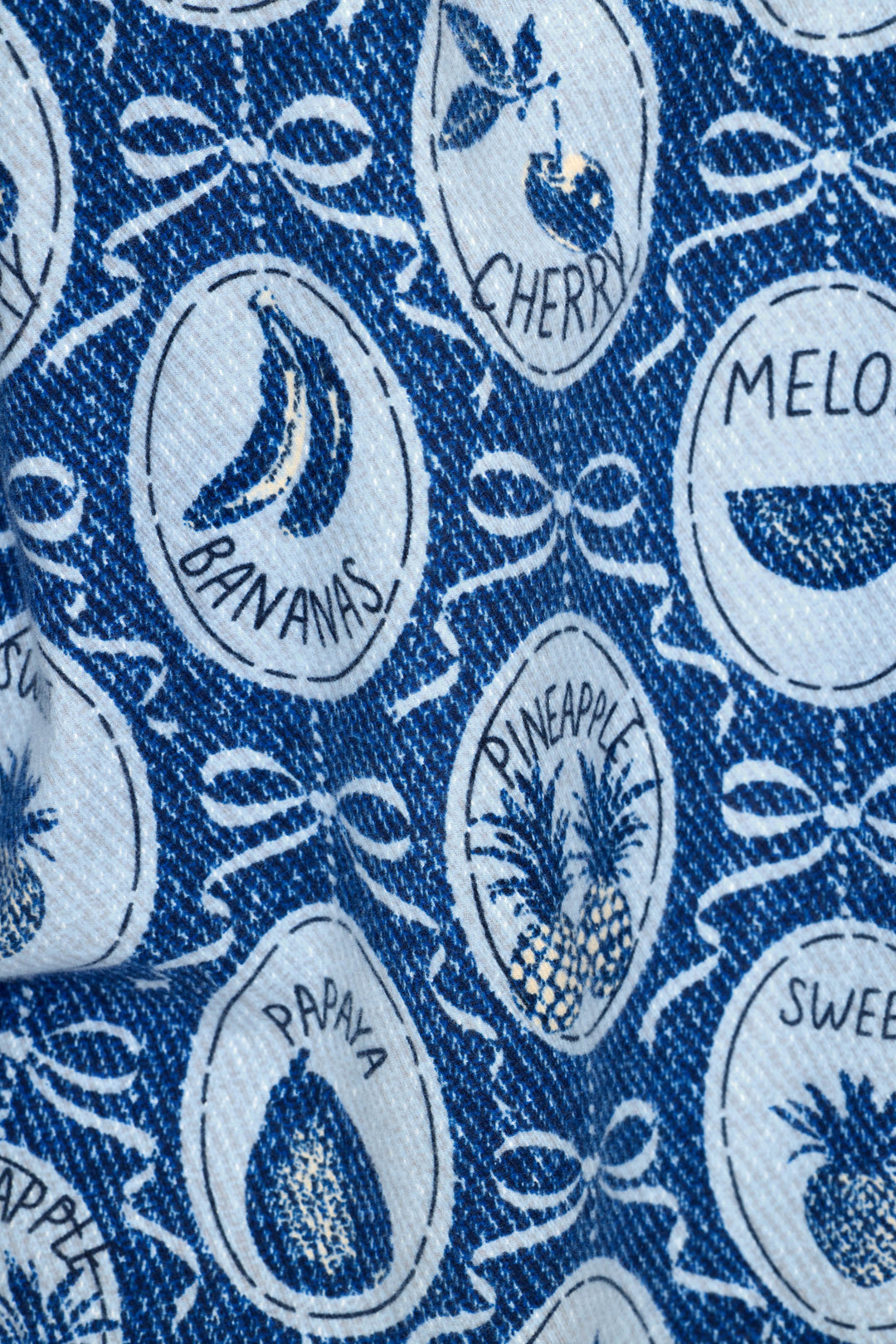 A close-up of a blue and white denim-like color fabric printed with white oval fruit illustrations and words like CHERRY, BANANAS, MELON, PINEAPPLE, PAPAYA, and SWEET, accents with bows.