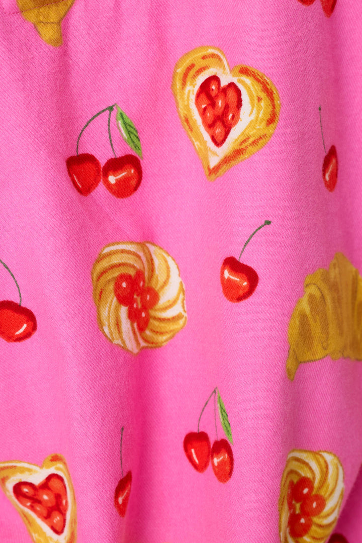 A close-up of a pink fabric with a repeating pattern of red cherries and heart-shaped pastry slices in yellow and cream.