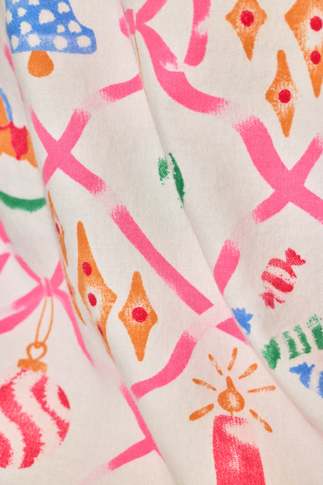 A close-up of a cream-colored fabric with a colorful, hand-painted style holiday pattern featuring pink ribbons, orange diamond ornaments, green accents, and red striped baubles.