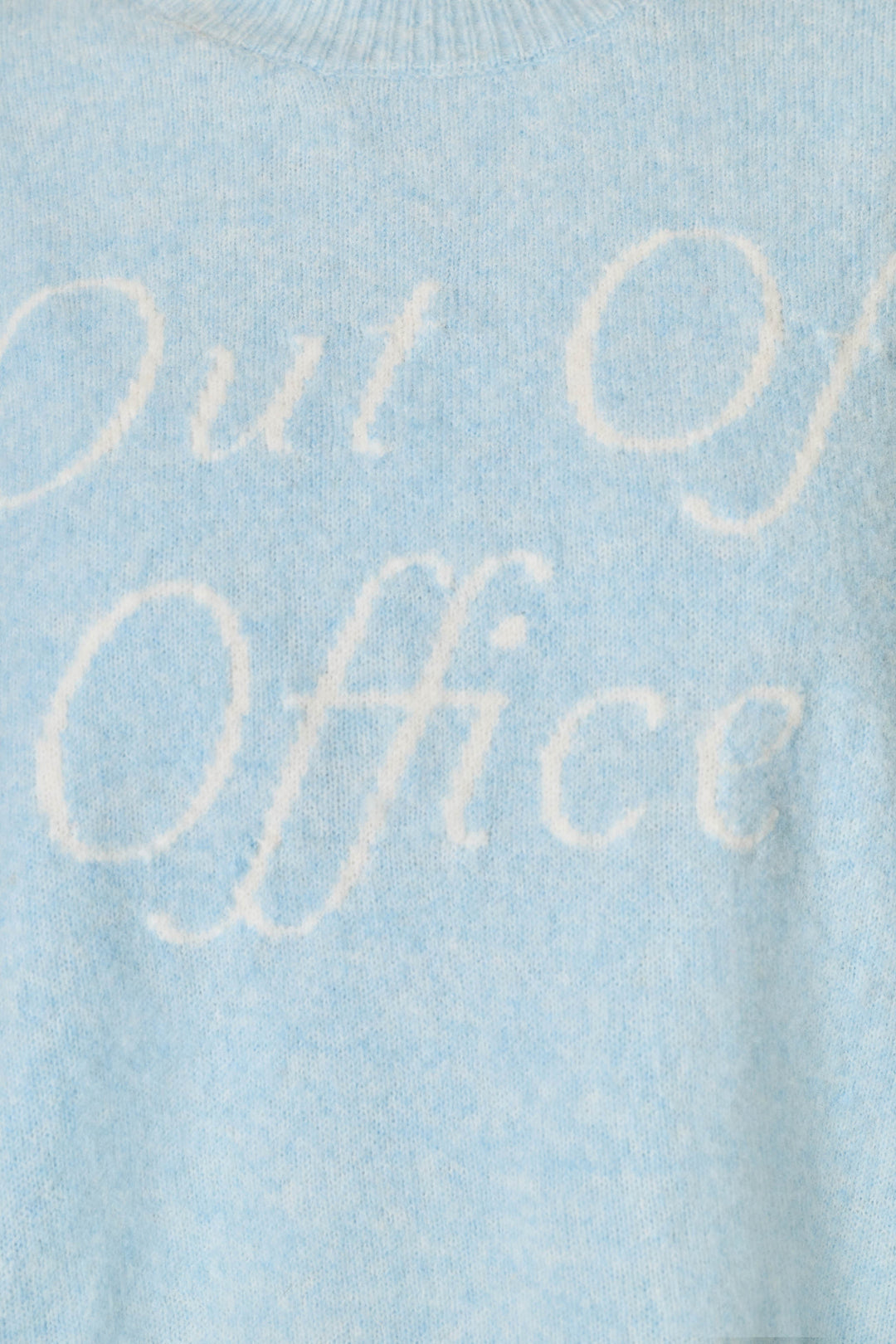 Close-up of a light blue knit sweater with the words 'Out of Office' knitted in a contrasting white script across the chest.