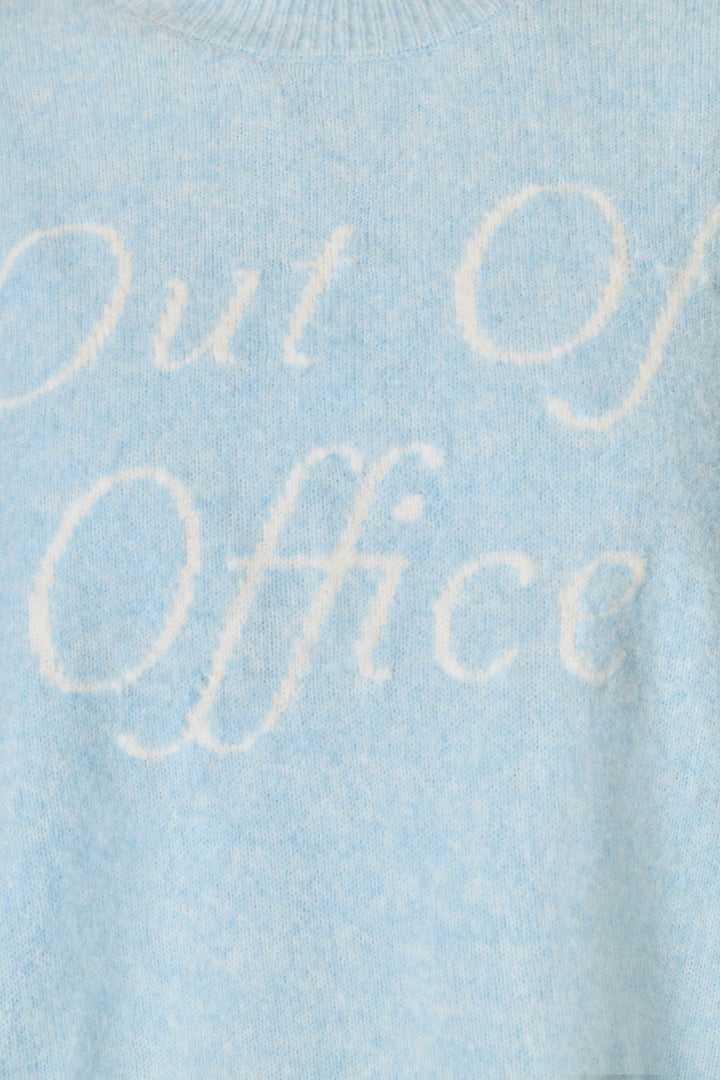 Close-up of a light blue knit sweater with the words 'Out of Office' knitted in a contrasting white script across the chest.