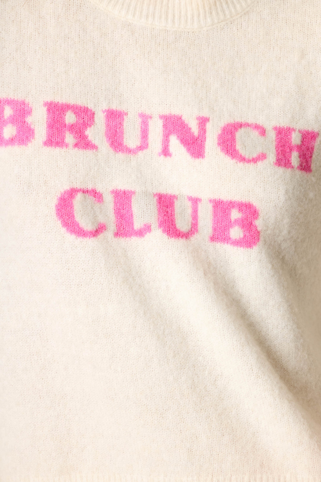 Close-up of a cream-colored knit sweater with the words 'BRUNCH CLUB' knitted in bright pink across the chest.