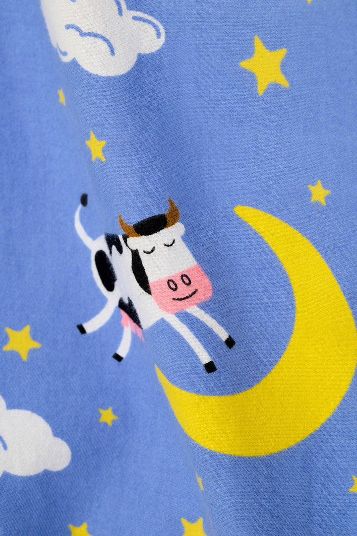 A close-up of a blue fabric with a playful cartoon print featuring a black-and-white cow with a pink snout jumping over a yellow crescent moon and small yellow stars, with white clouds also visible.