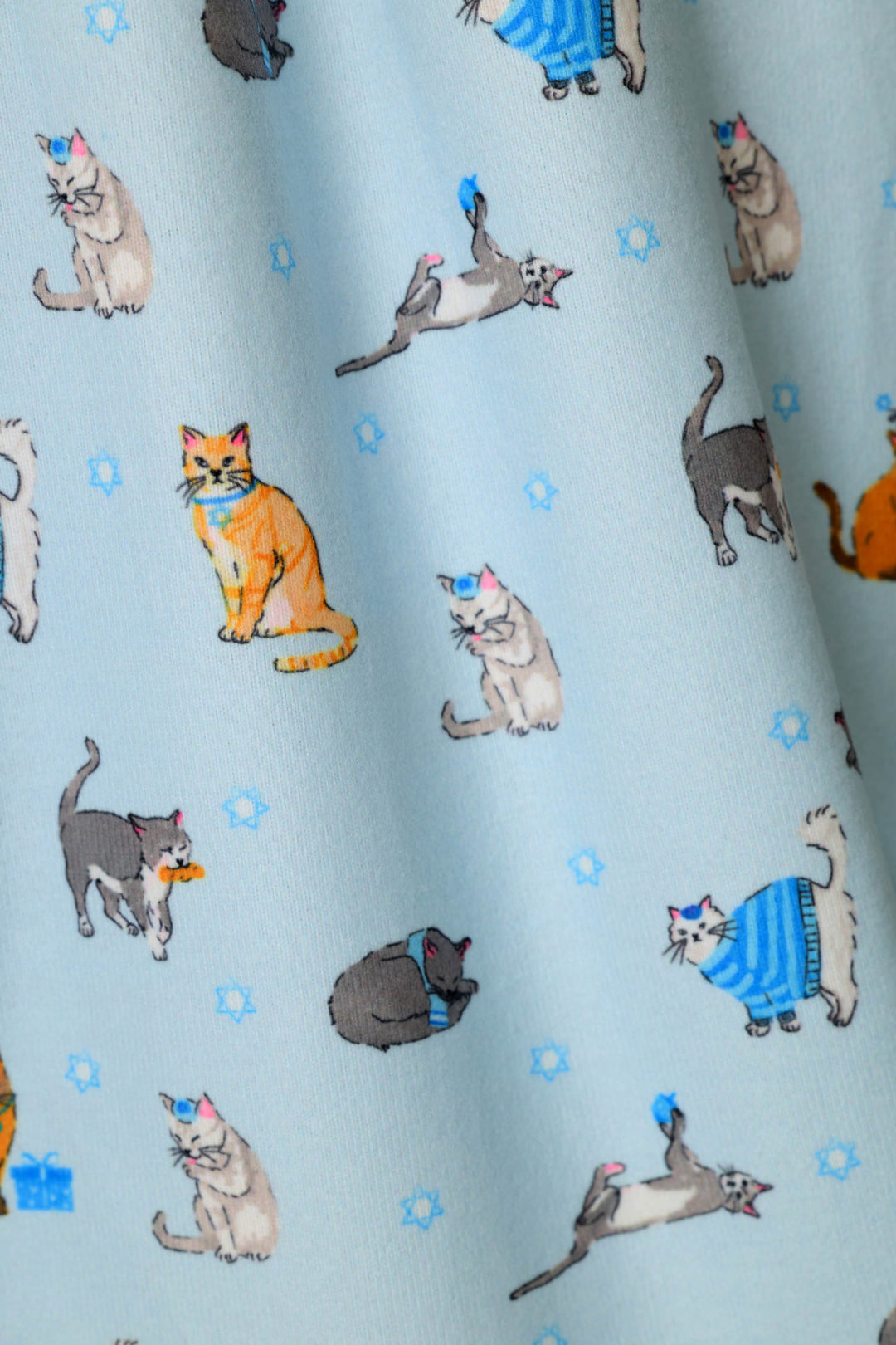 Light blue fabric printed with a playful pattern of cats in various poses, including some wearing blue striped sweaters, playing with dreidels, or wearing yarmulkas. 