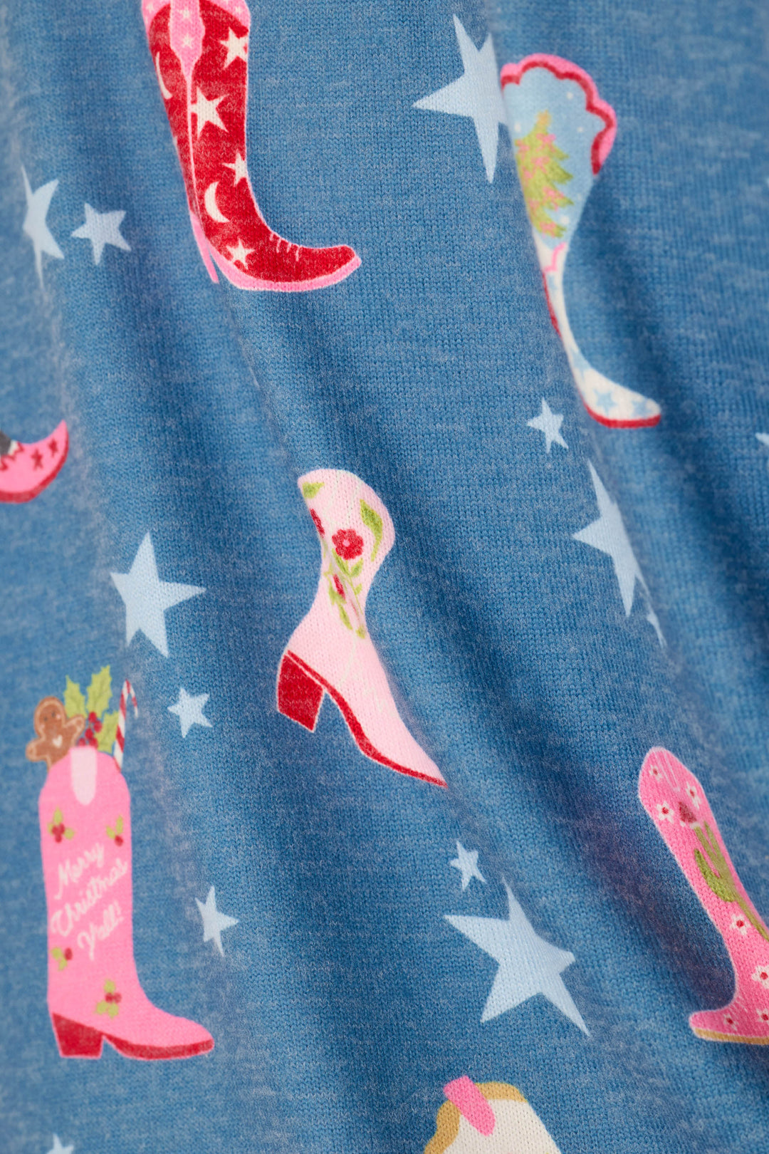 A close-up of a blue fabric surface printed with cowboy boots and white stars. The boots are in pink, red, and white, with decorative floral and holiday motifs.