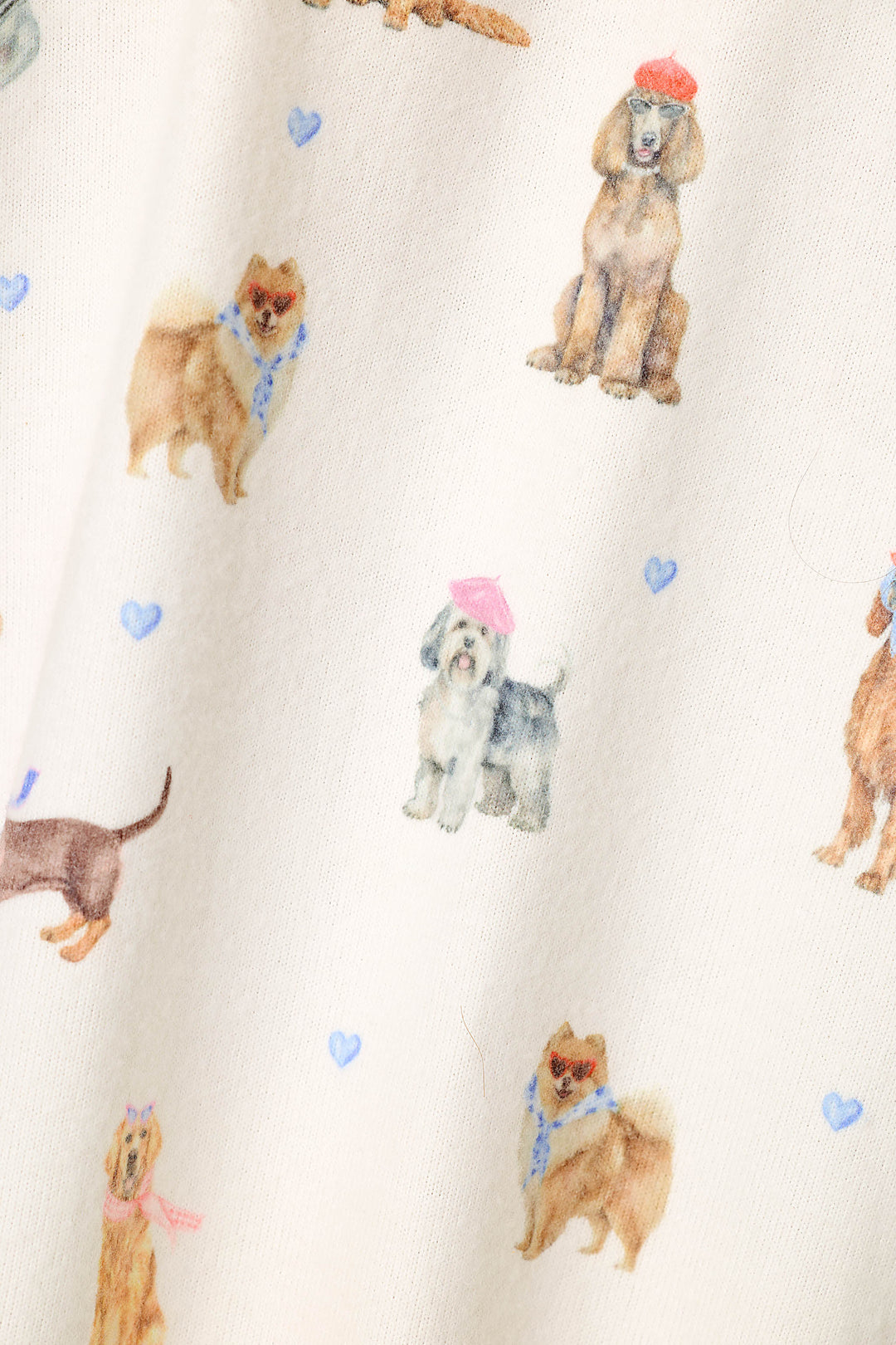 Close-up of a cream-colored fabric with a watercolor print of various dog breeds, including Pomeranians, Poodles, and Shih Tzu dogs, some wearing pink and red hats and blue scarves, accented by small blue hearts.