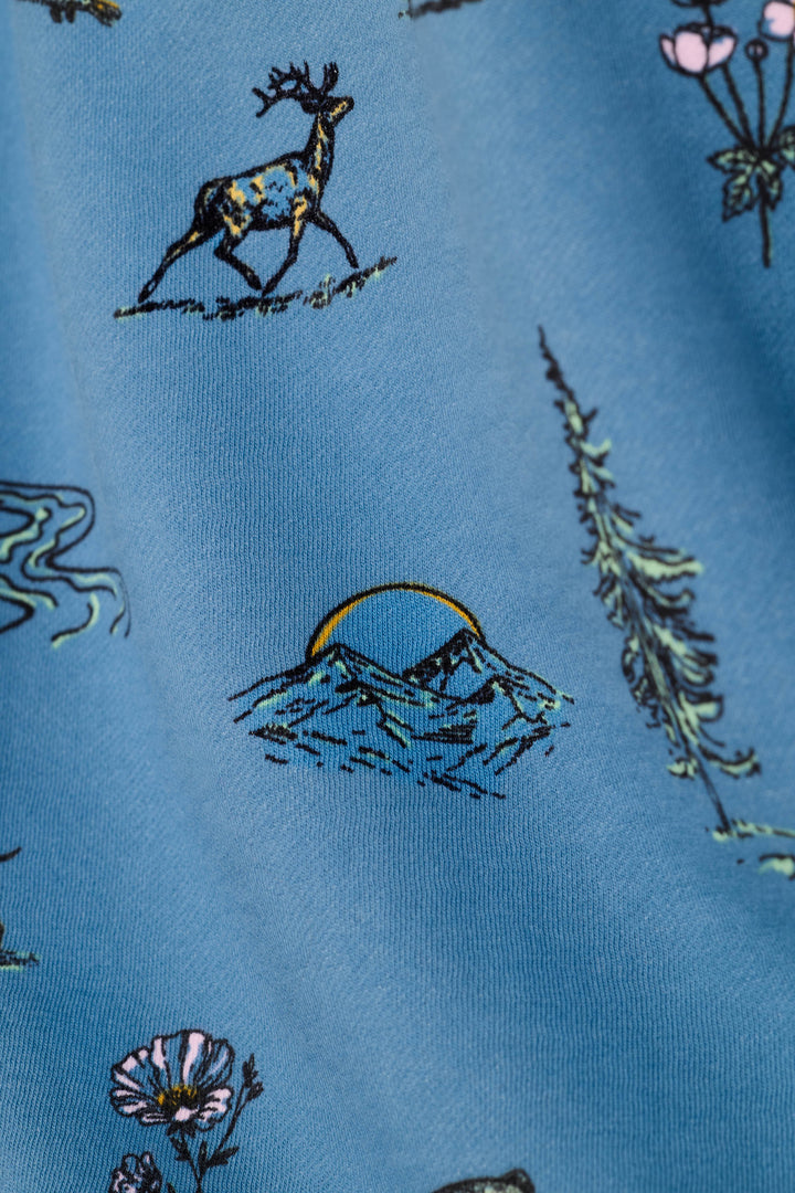A close-up of a blue fabric featuring a repeating pattern of hand-drawn nature illustrations, including a deer, mountains with a rising sun, pine trees, and wildflowers.