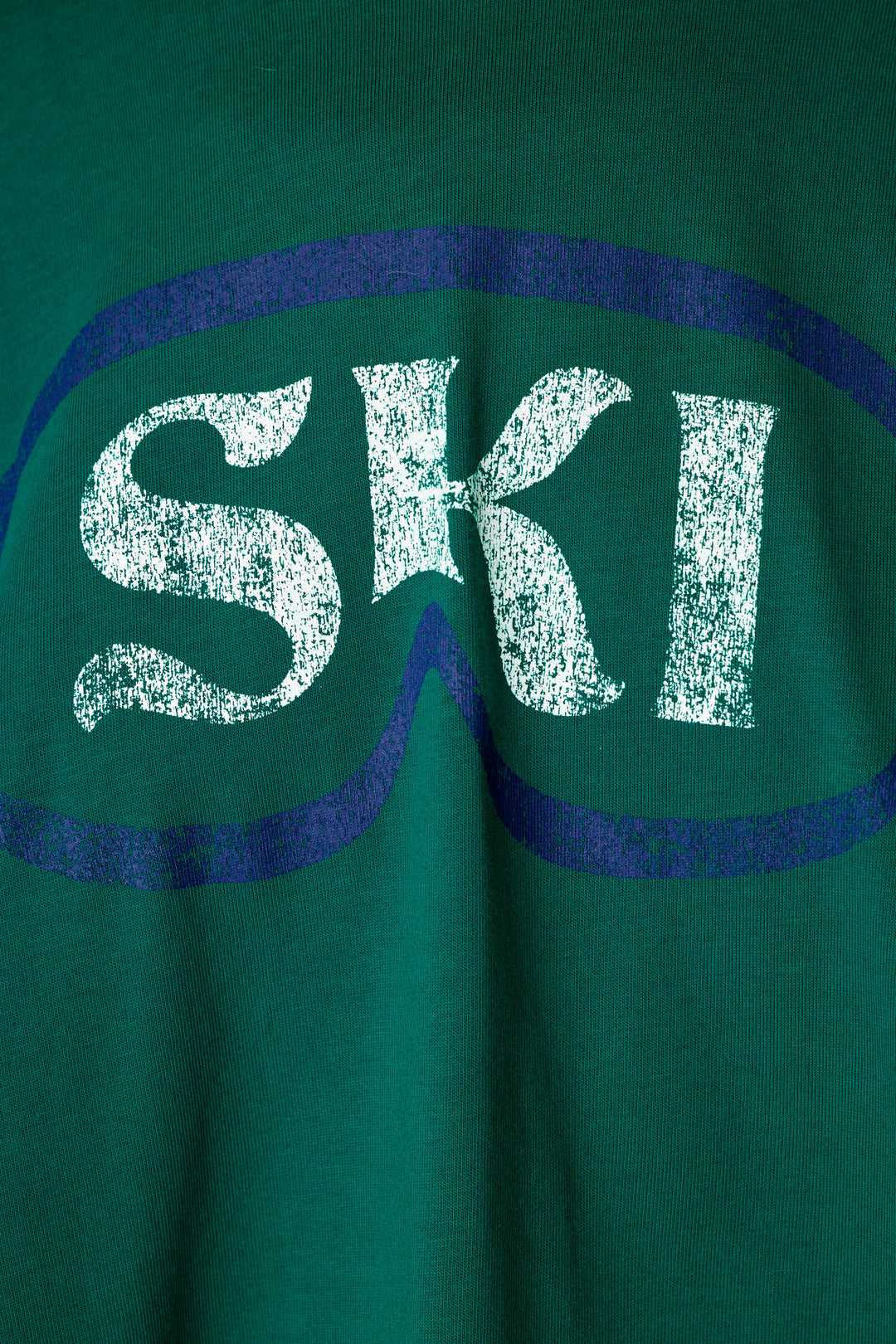 Close-up of a green knit garment featuring a distressed white 'SKI' graphic inside a dark blue ski goggles outline.