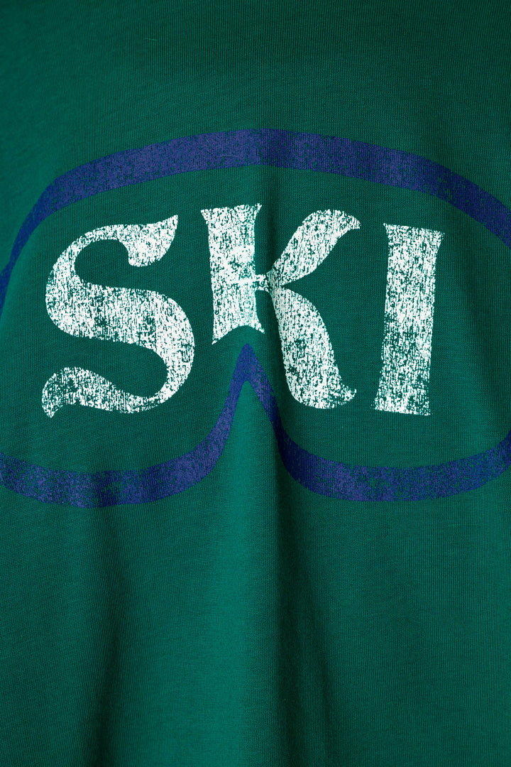 Close-up of a green knit garment featuring a distressed white 'SKI' graphic inside a dark blue ski goggles outline.