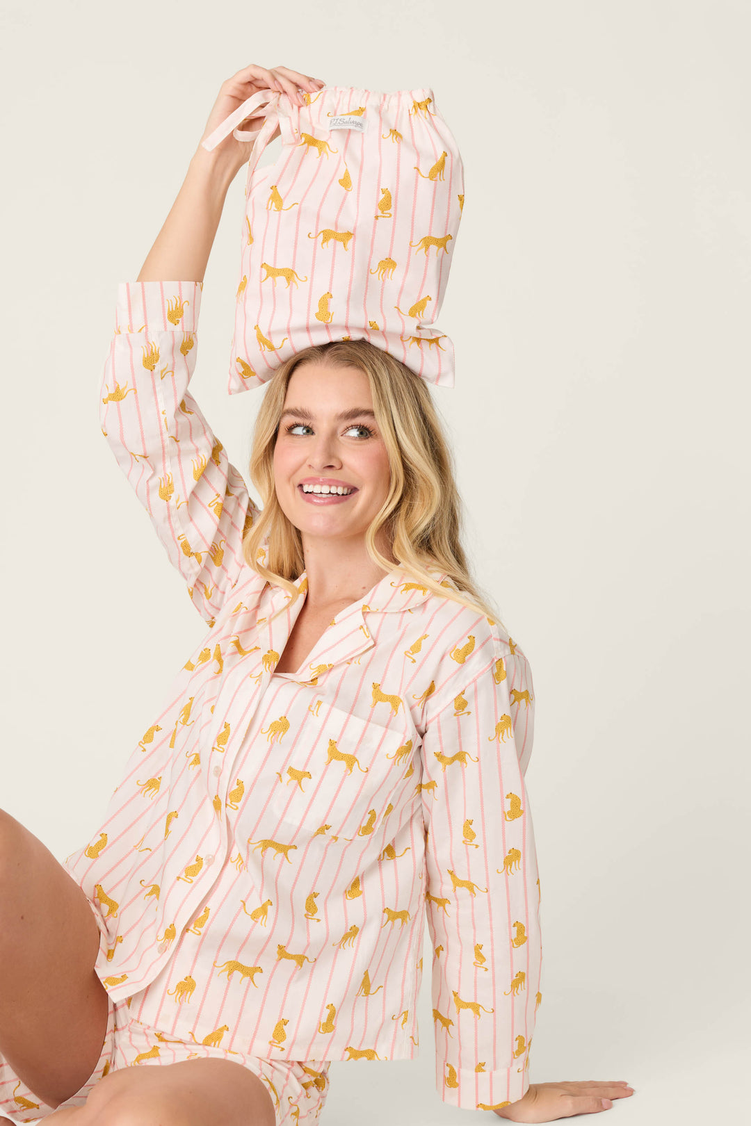 A person wearing the P.J. Salvage Afterparty Poplin Cheetah Charm Short PJ Set balances a matching drawstring bag on their head and smiles against a plain light background.