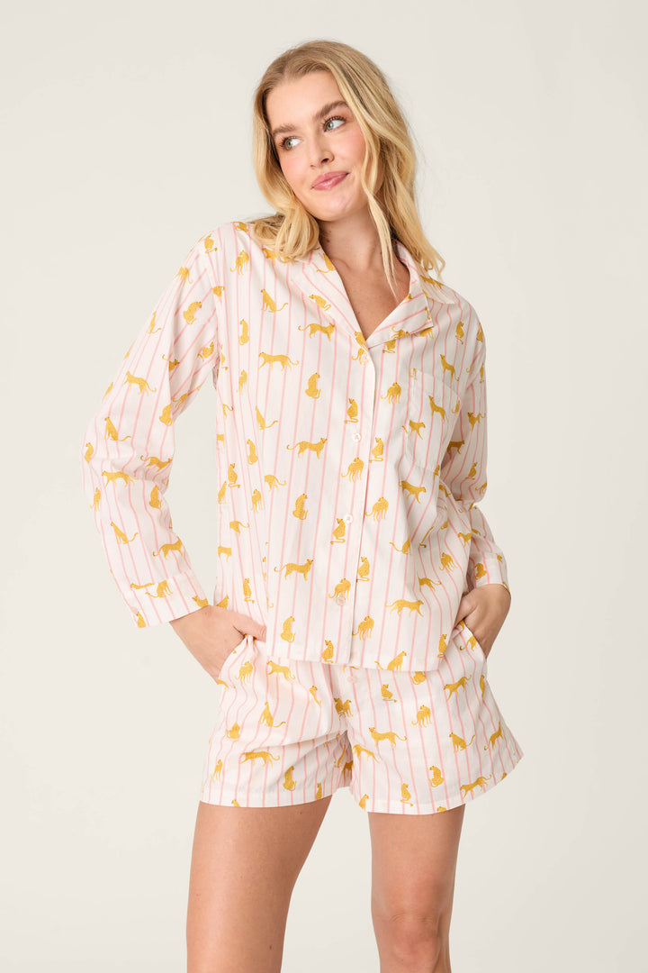 A person wears the P.J. Salvage Afterparty Poplin Cheetah Charm Short PJ Set, featuring a button-up long-sleeve top and shorts in light pink and white stripes with small yellow designs, against a plain off-white background.