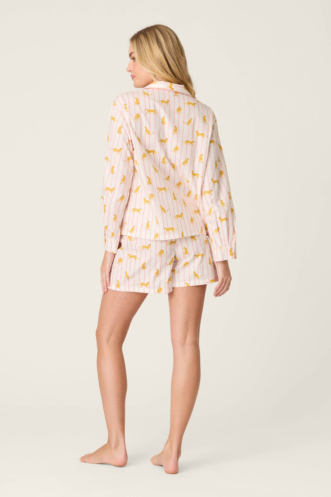 A person stands barefoot on a light background, facing away from the camera, wearing the P.J. Salvage Afterparty Poplin Cheetah Charm Short PJ Set—a pink pajama set with a long-sleeve top and shorts in playful yellow cheetah print.
