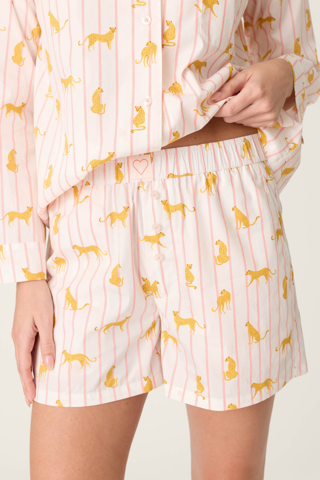 A person wears the Afterparty Poplin Cheetah Charm Short PJ Set by P.J. Salvage, featuring a pink cheetah print with white stripes and yellow accents. The image highlights the lower torso, one hand lifting the shirt to show matching shorts.