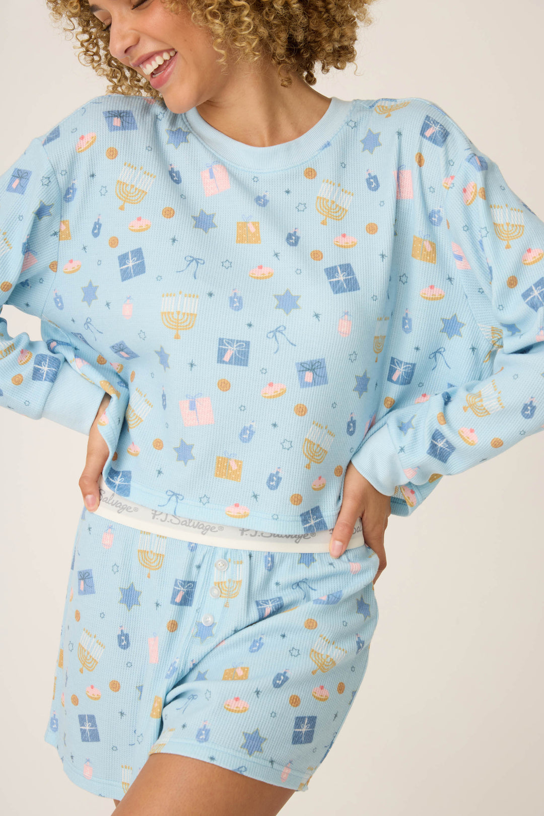 A person wears the P.J. Salvage STARS PJ SET BOYFRIEND BOXER SHORT GIFT SETS, a light blue Hanukkah-themed pajama set featuring menorahs, dreidels, gifts, and stars. They smile with one hand on their hip.