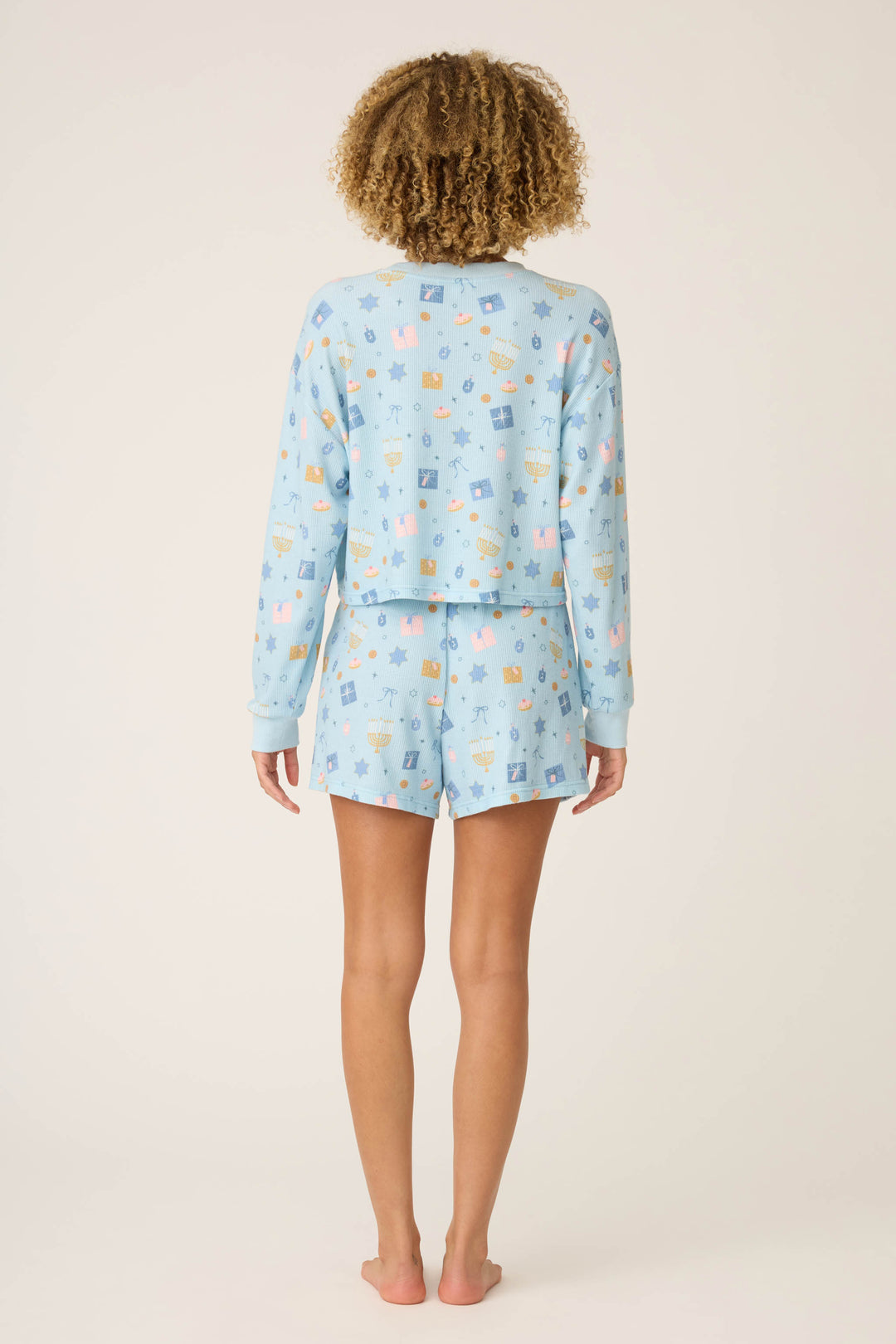 A barefoot person with curly hair faces away, wearing the P.J. Salvage STARS PJ SET BOYFRIEND BOXER SHORT GIFT SETS—light blue long-sleeve top and matching cartoon-print shorts—against a plain backdrop.