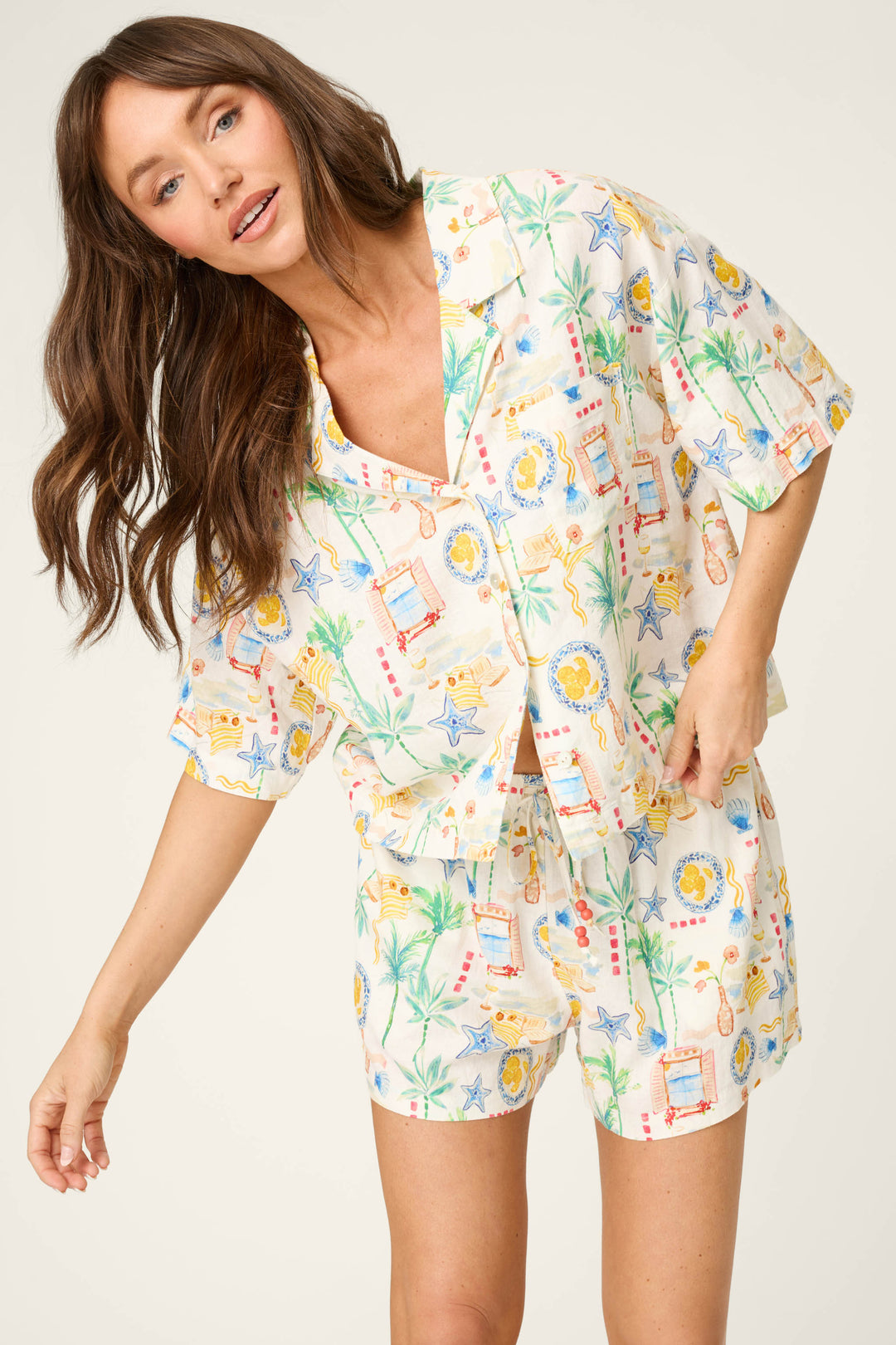 A woman with long brown hair models a playful short-sleeve button-up shirt and shorts with a colorful print on a light background.