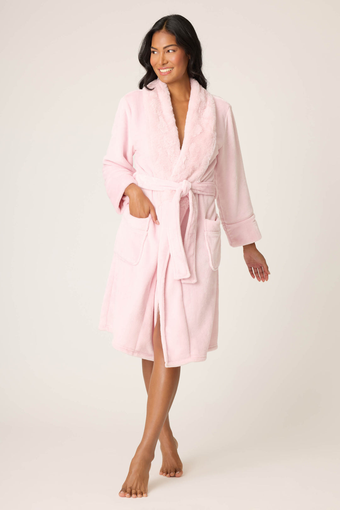 A person, barefoot on a neutral background, wears the P.J. Salvage Luxe Plush Solid Pink Robe featuring a soft plush texture, shawl collar, belted waist, and two front pockets. They look to the side and smile.