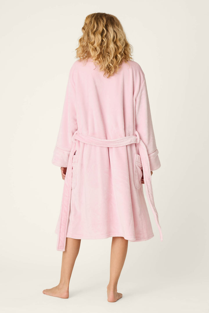 A person with shoulder-length curly blond hair, seen from behind, is standing barefoot on a plain light background, wearing the P.J. Salvage Luxe Plush Solid Pink Robe—a mid-length robe featuring plush fleece fabric and a tied belt.
