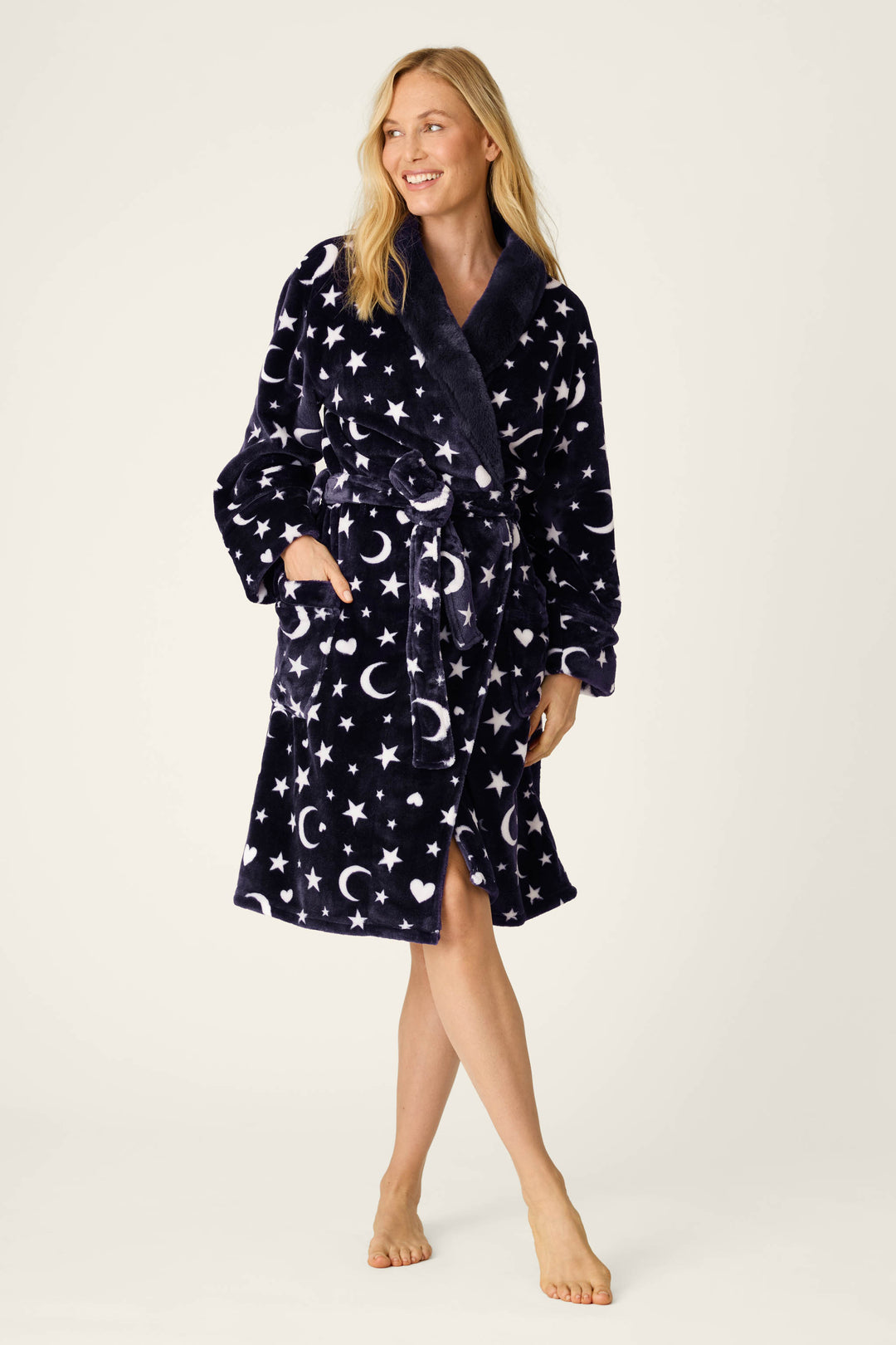 A woman stands barefoot, smiling and looking to the side, wearing the P.J. Salvage Luxe Plush Celestial Print Robe in navy blue with white star and moon patterns; one hand is tucked into her pocket while the other rests by her side.
