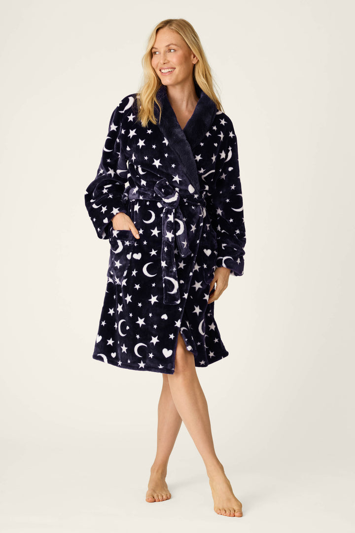 A woman stands barefoot, smiling and looking to the side, wearing the P.J. Salvage Luxe Plush Celestial Print Robe in navy blue with white star and moon patterns; one hand is tucked into her pocket while the other rests by her side.
