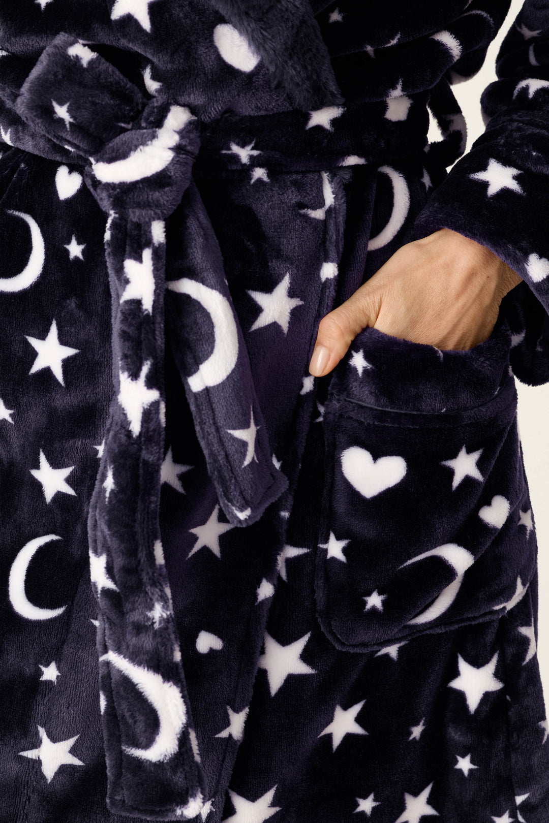 Close-up of a plush robe in dark navy with a white celestial print of stars, moons, and hearts, featuring a side pocket and a hand reaching into the pocket.