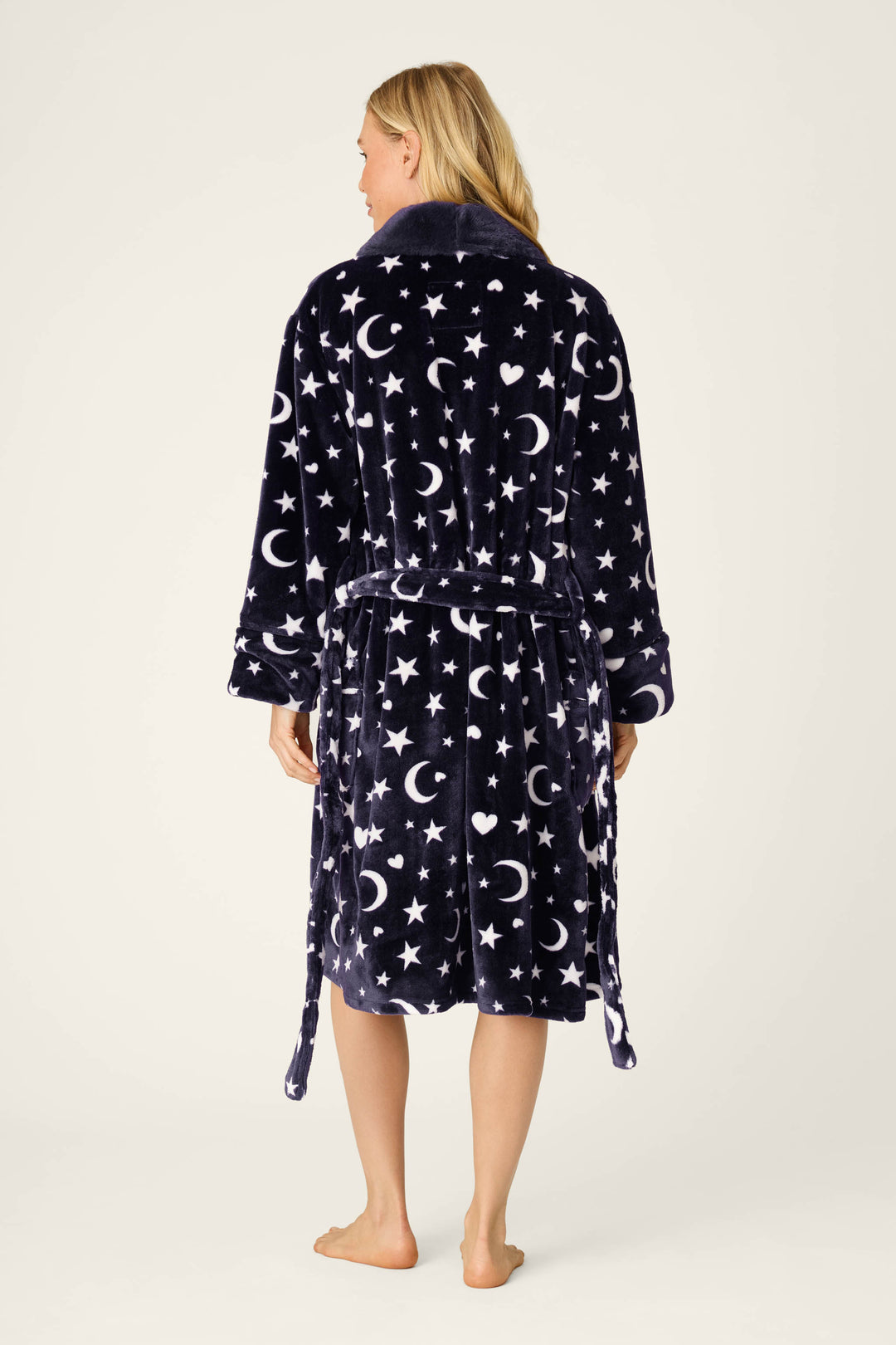 A person stands barefoot, facing away, wearing the P.J. Salvage Luxe Plush Celestial Print Robe featuring a dark plush fabric with white crescent moon, star, and heart designs. The robe includes long sleeves and a belt tied at the waist.
