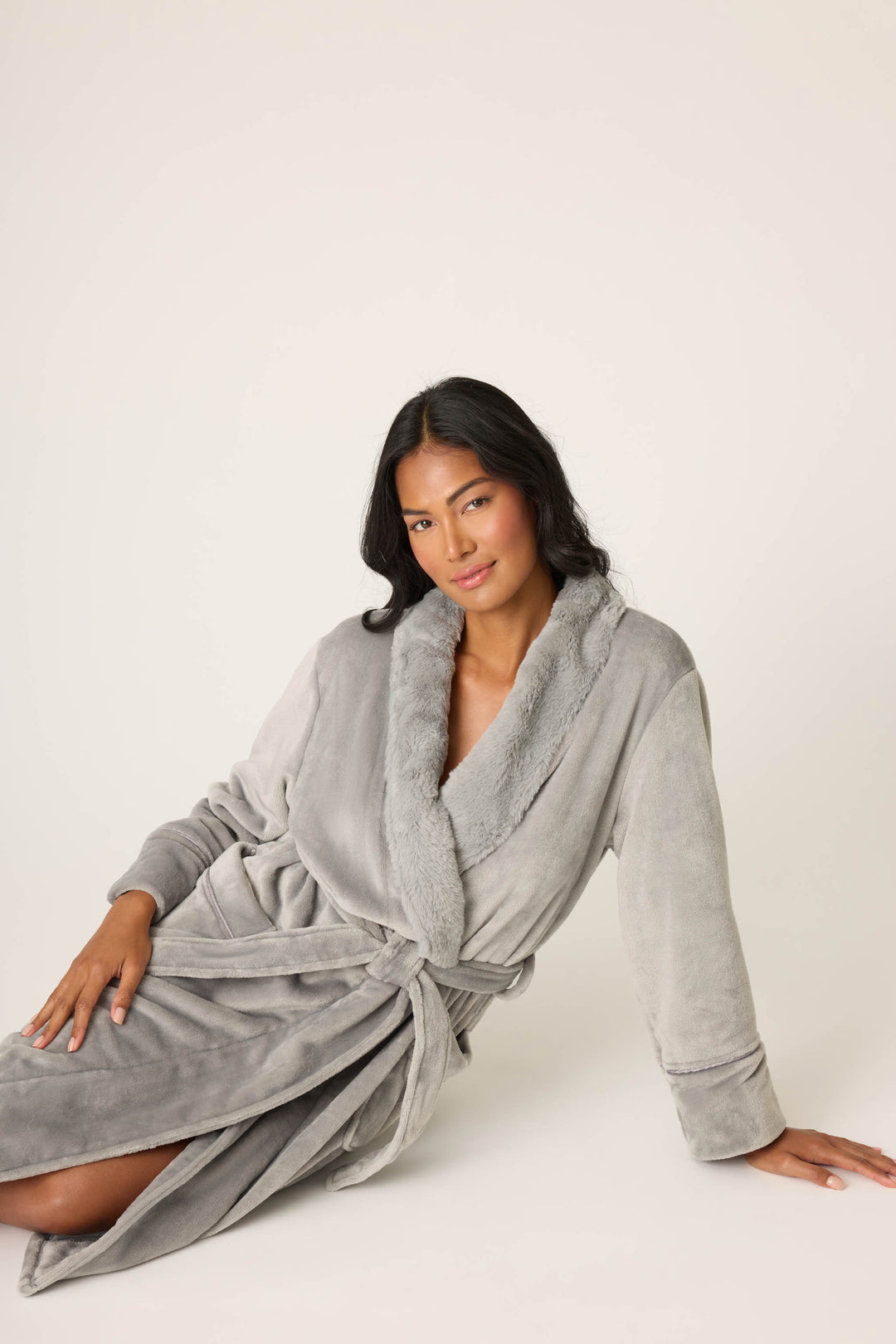 A woman with long dark hair sits on the floor against a plain off-white background, wearing the P.J. Salvage Luxe Plush Solid Frost Gray Robe featuring a tie belt and shawl collar.