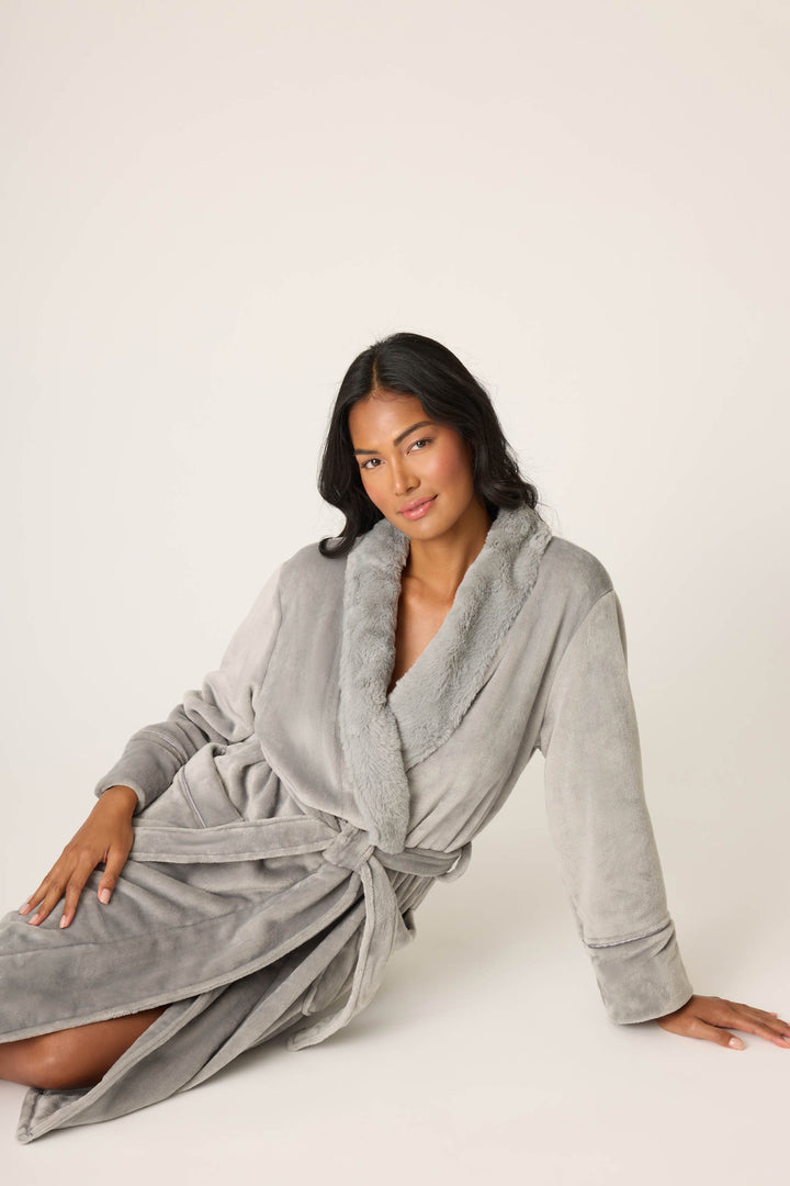 A woman with long dark hair sits on the floor against a plain off-white background, wearing the P.J. Salvage Luxe Plush Solid Frost Gray Robe featuring a tie belt and shawl collar.