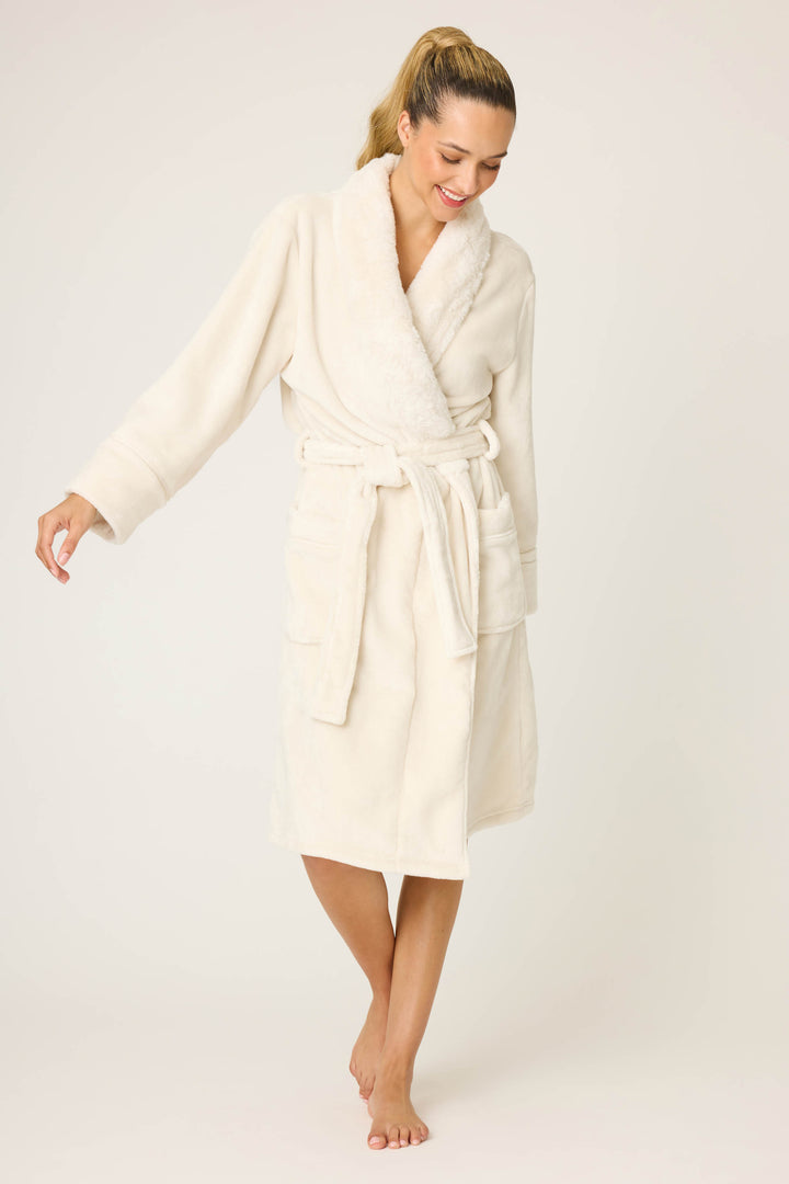 A woman models the P.J. Salvage Luxe Plush Solid Ivory Robe, featuring a knee-length fit, tie belt, and pockets. She stands barefoot against a light background, smiling and looking down with one foot in front of the other.