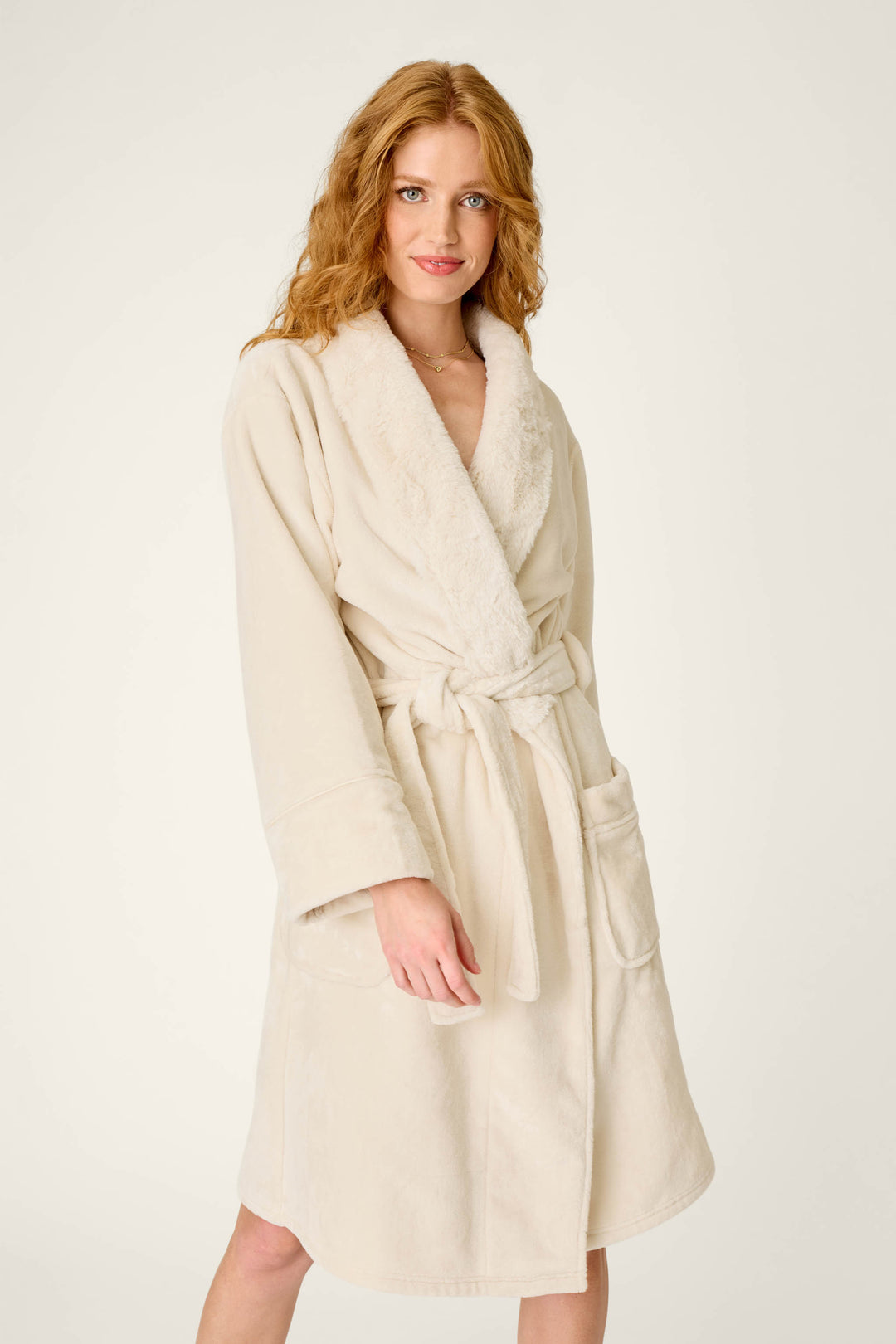 A woman with long, wavy red hair is wearing the P.J. Salvage Luxe Plush Solid Natural Robe, a silky-soft fleece robe featuring a shawl collar, tie belt, and front pockets, standing against a plain light background.