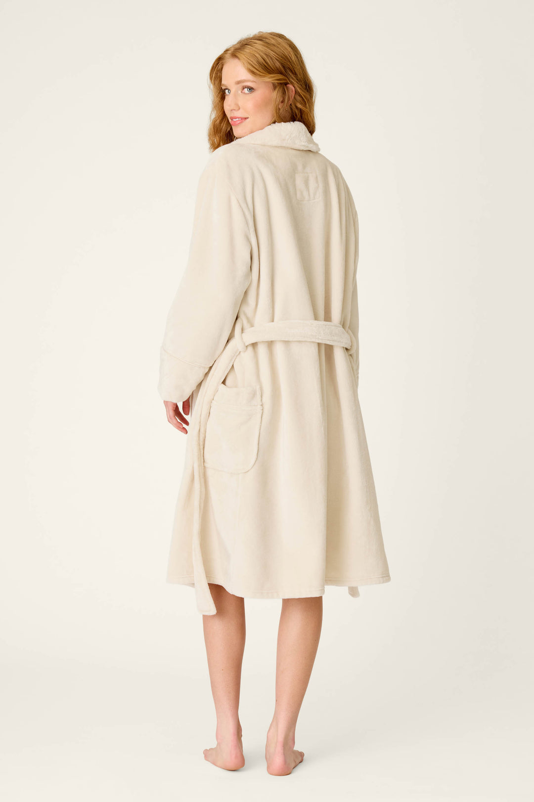 A woman with long hair stands barefoot, facing away and glancing back over her shoulder with a slight smile, wearing the P.J. Salvage Luxe Plush Solid Natural Robe featuring a silky-soft fleece texture and a shawl collar.