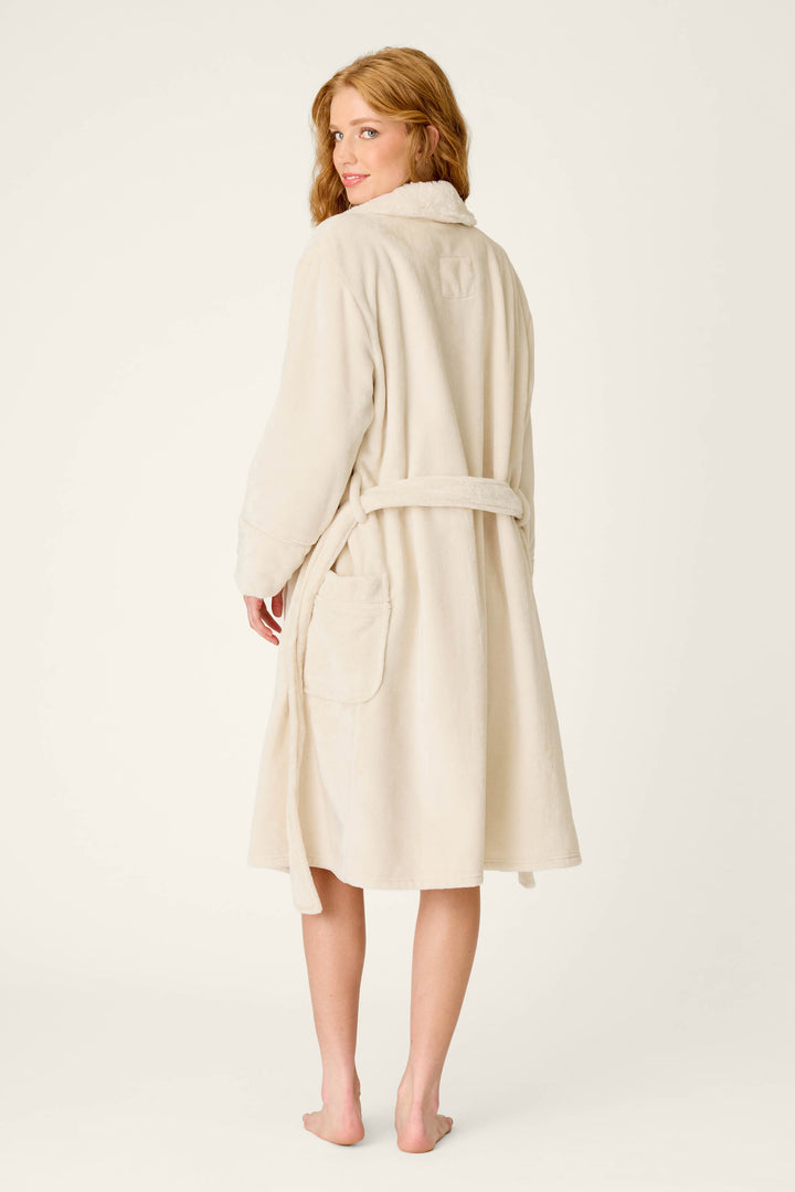 A woman with long hair stands barefoot, facing away and glancing back over her shoulder with a slight smile, wearing the P.J. Salvage Luxe Plush Solid Natural Robe featuring a silky-soft fleece texture and a shawl collar.
