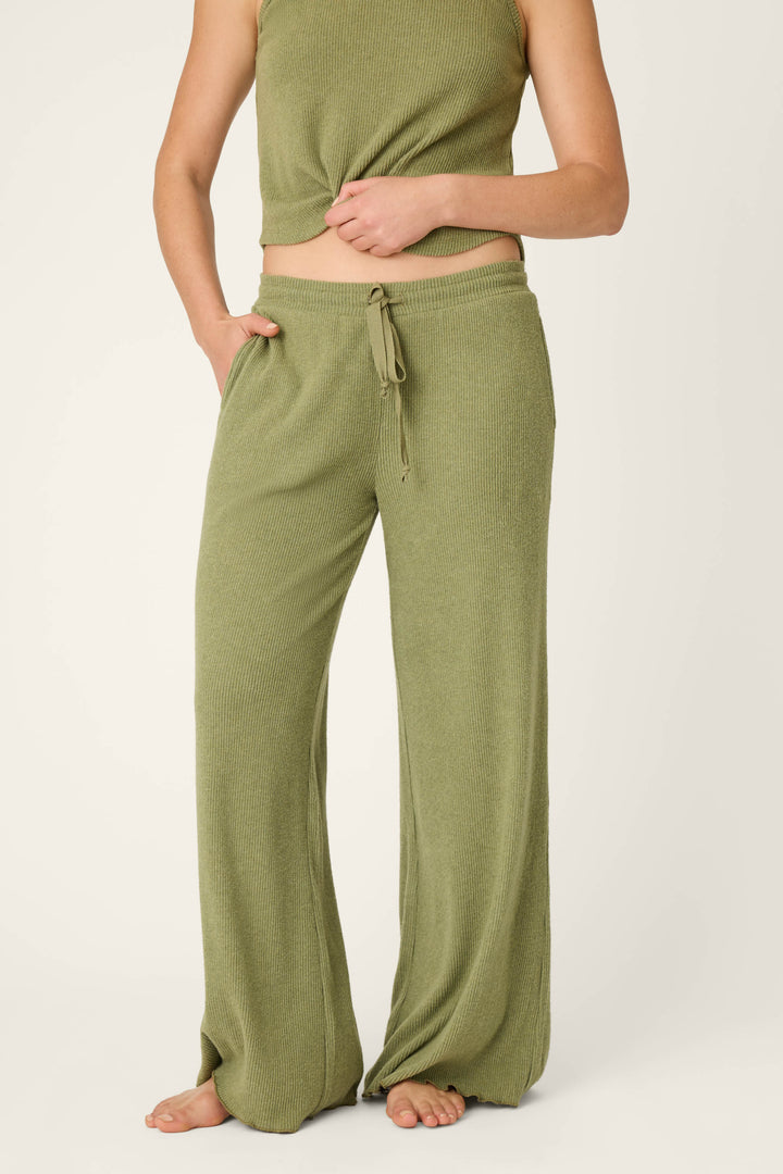 A person stands barefoot wearing P.J. Salvage Olive You Forever Lounge Pants—loose, olive green ribbed knit with a drawstring waist—paired with a matching sleeveless top, partially lifting the shirt to reveal the fit of the pants.