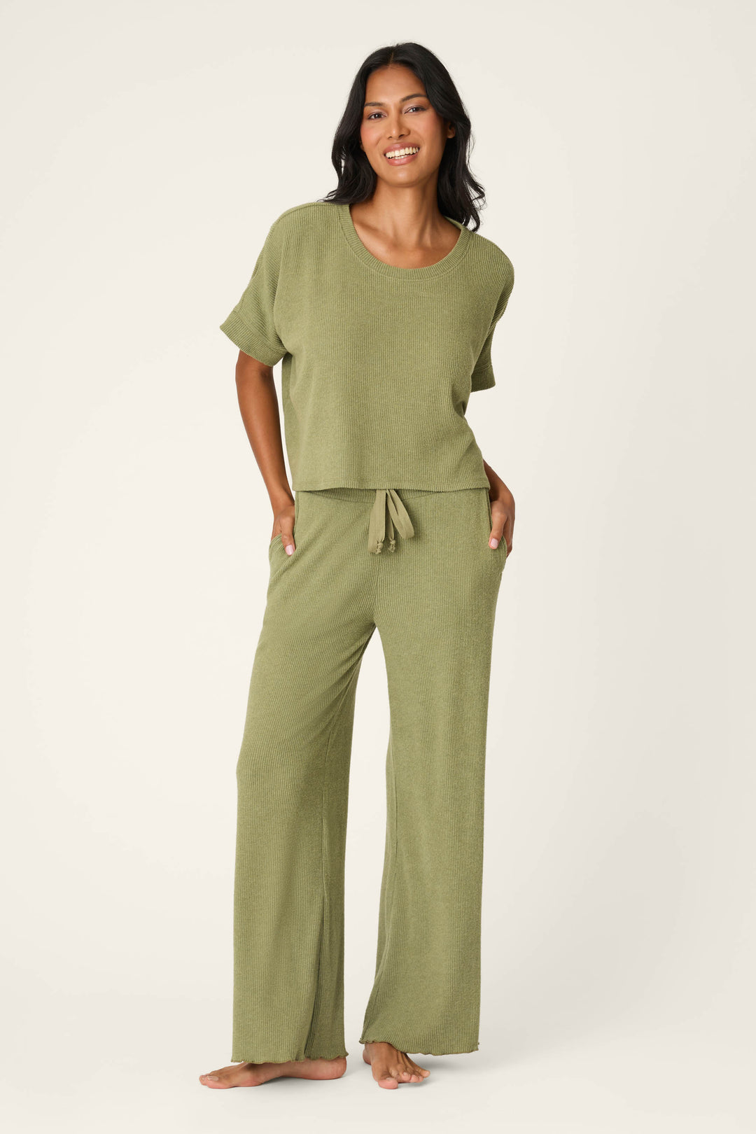 A woman smiles barefoot in the Olive You Forever Lounge Set by P.J. Salvage—an olive green, loose-fitting short-sleeve top with matching wide-leg, drawstring pants—posed against a plain light background.
