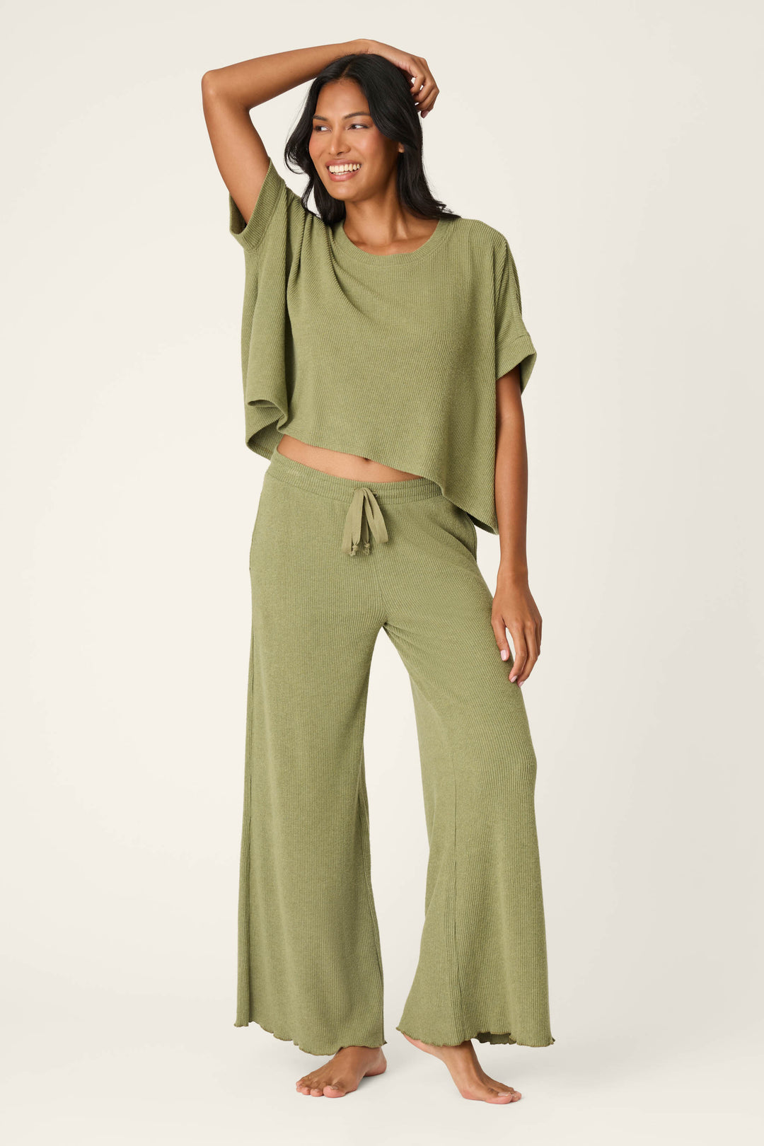 A woman smiles and poses barefoot, wearing the P.J. Salvage Olive You Forever Lounge Set—an olive green short-sleeve top and matching wide-leg drawstring pants—against a plain light background.
