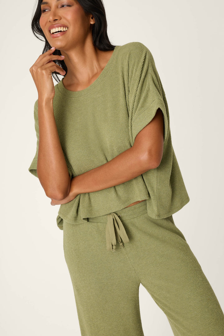 A woman smiles in the P.J. Salvage Olive You Forever Lounge Set—a loose-fitting, textured olive green outfit with a short-sleeve top and matching drawstring pants—standing against a plain light background.